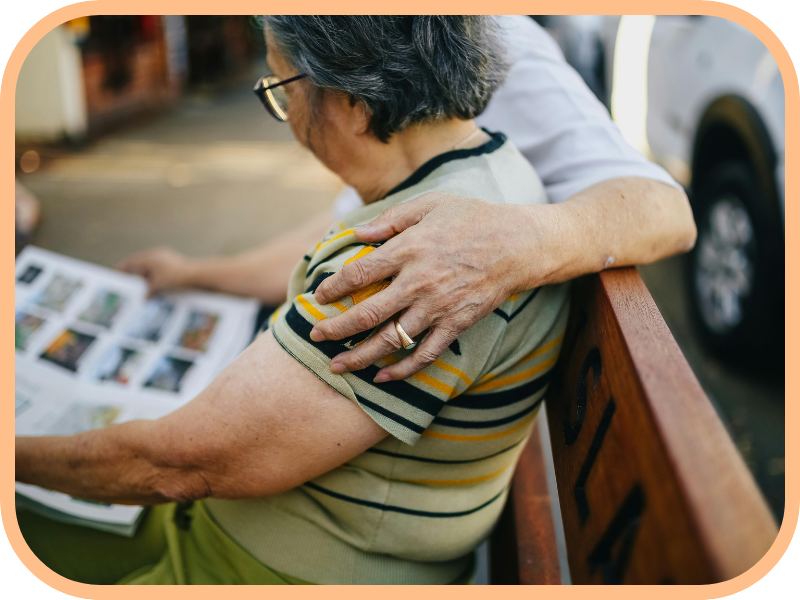 Caregiver rests a hand on an older woman’s shoulder while she looks at photos outdoors