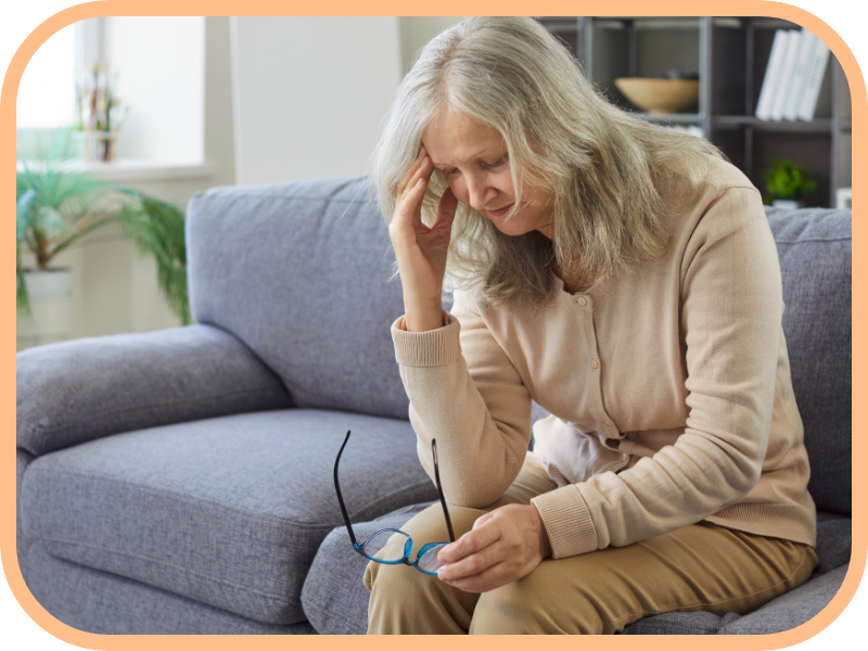 Older woman sits on a couch holding glasses and resting her head at home