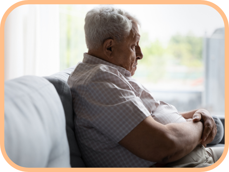 Older man sits quietly on a couch at home, looking out a window
