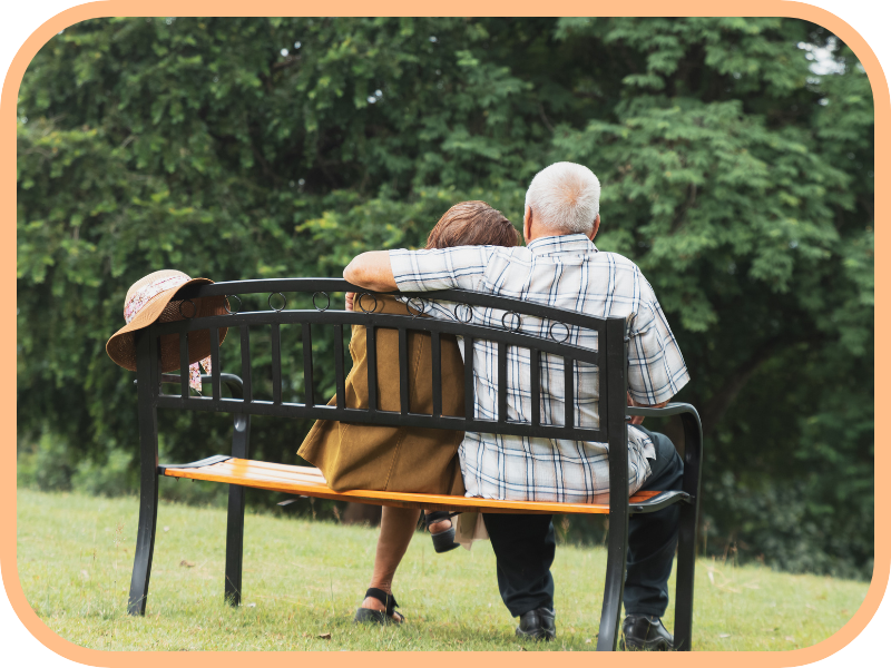 Older couple sits together on a park bench with arms around each other