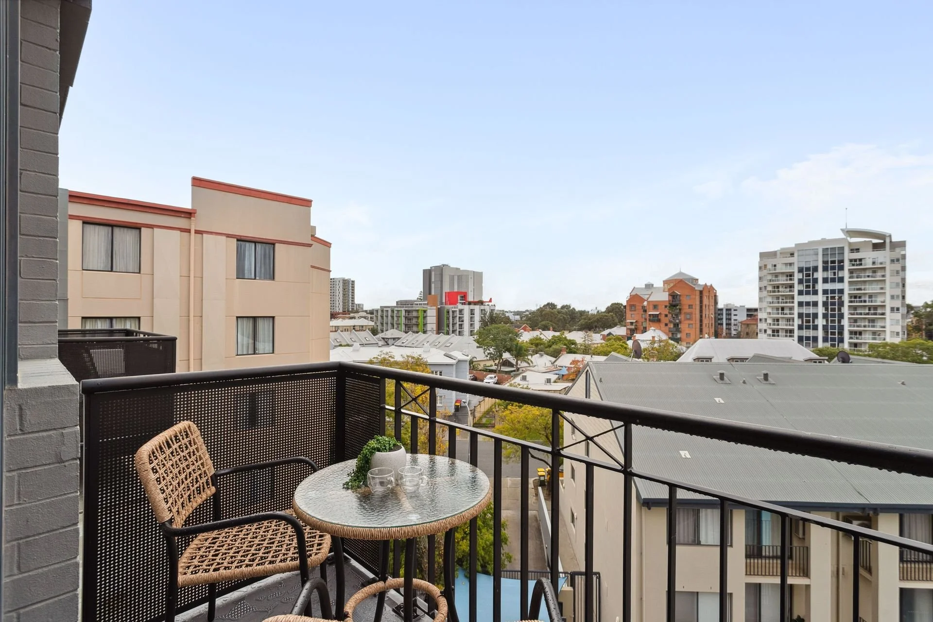 Balcony with a glass-topped table, a wicker chair, and a potted plant, overlooking a cityscape with various apartment buildings and trees under a clear sky.