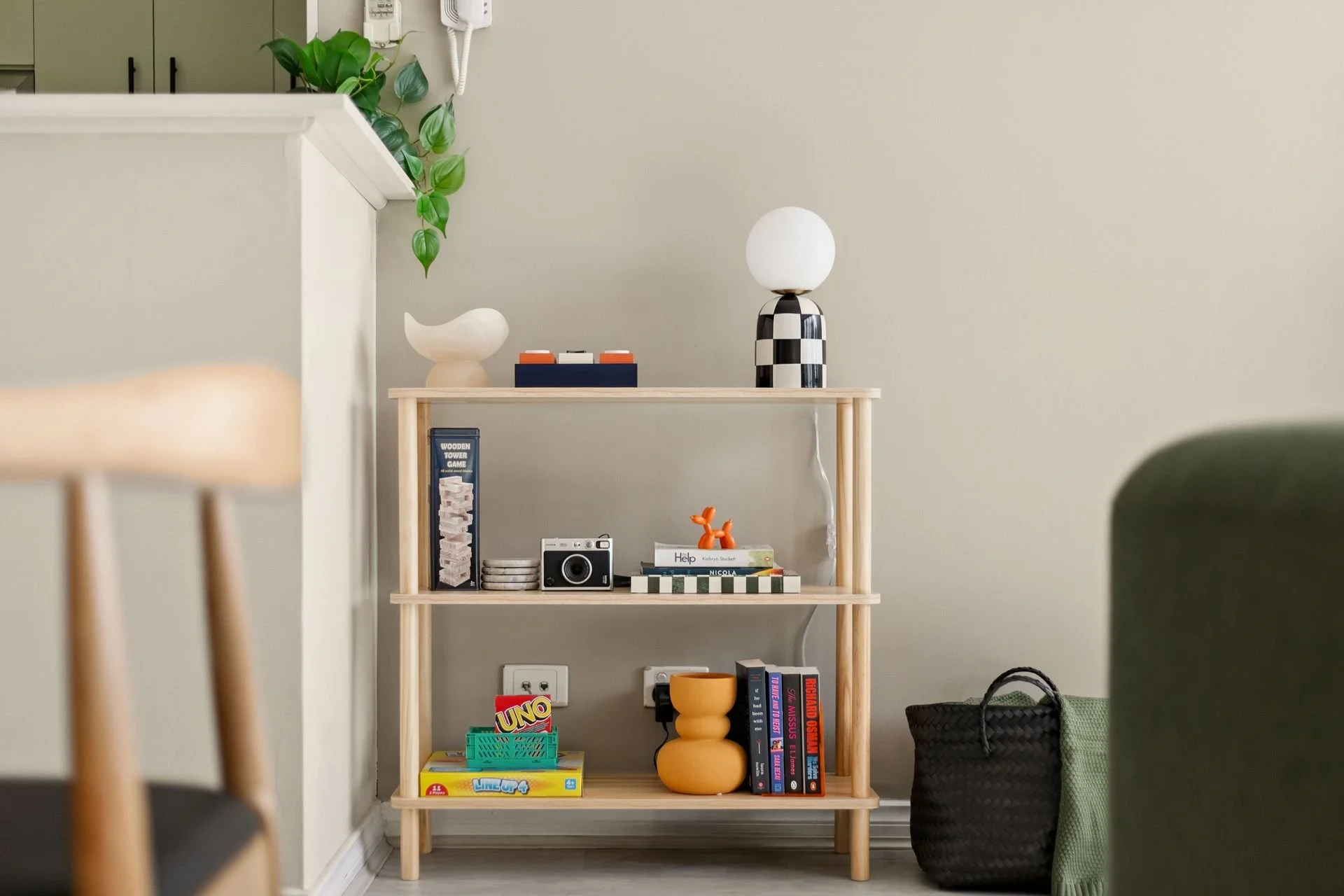 A wooden shelf with decorative items including a black and white checkered lamp with a white globe, a white ceramic bird, and a small orange sculpture. The shelf also holds books, a vintage camera, and a board game. To the right of the shelf are a gr