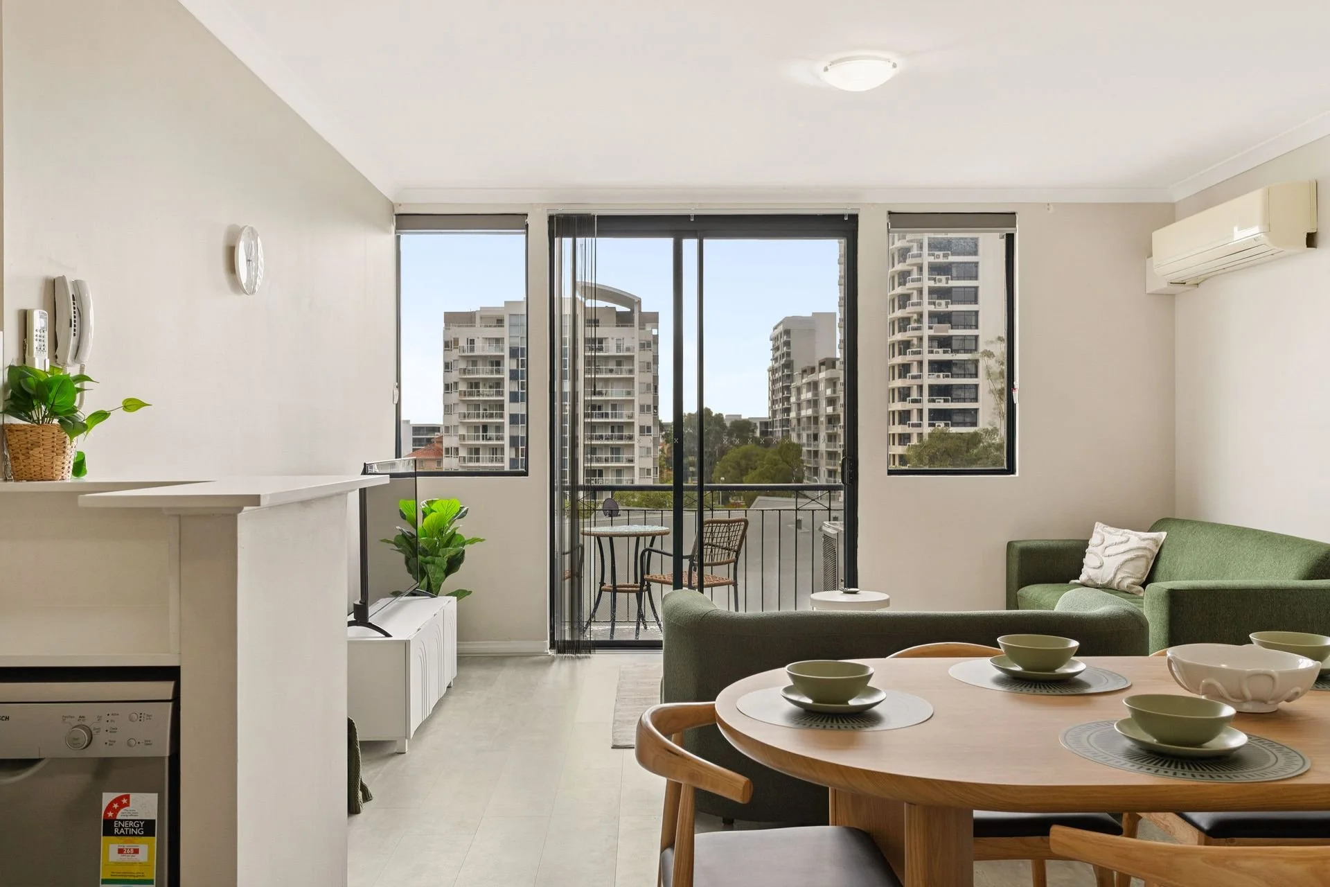 Living room with a green sofa, a round dining table with bowls, a balcony with outdoor chairs, and city buildings outside.
