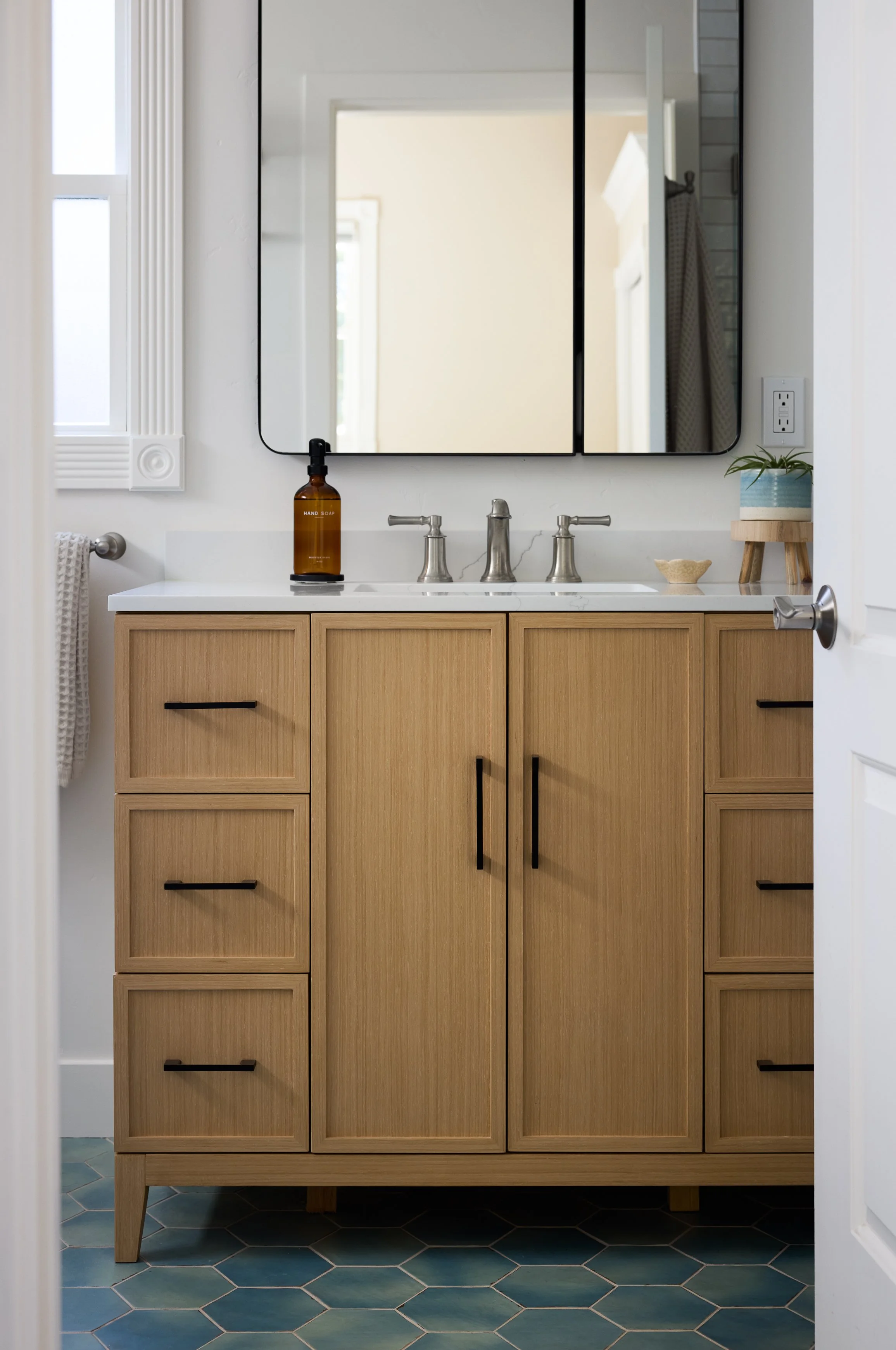 Transitional, coastal bathroom designed by Kelsey Brown Interiors in Aptos, California. Featuring a light oak vanity with black accents and a brushed nickel faucet. Blue green hexagon tiles as the bathroom flooring. 