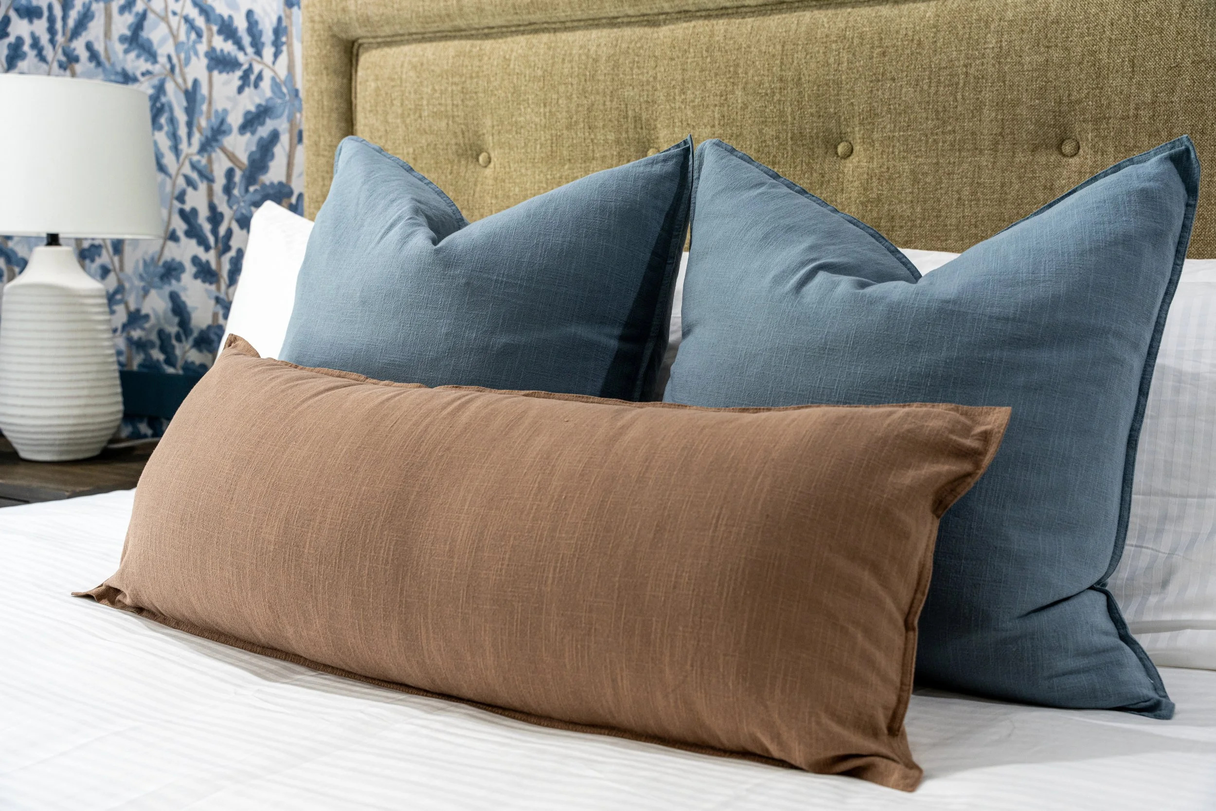 A close up of a bed with blue and brown throw pillows with a blue floral wallpaper in Asheville, North Carolina by Kelsey Brown Interiors. 