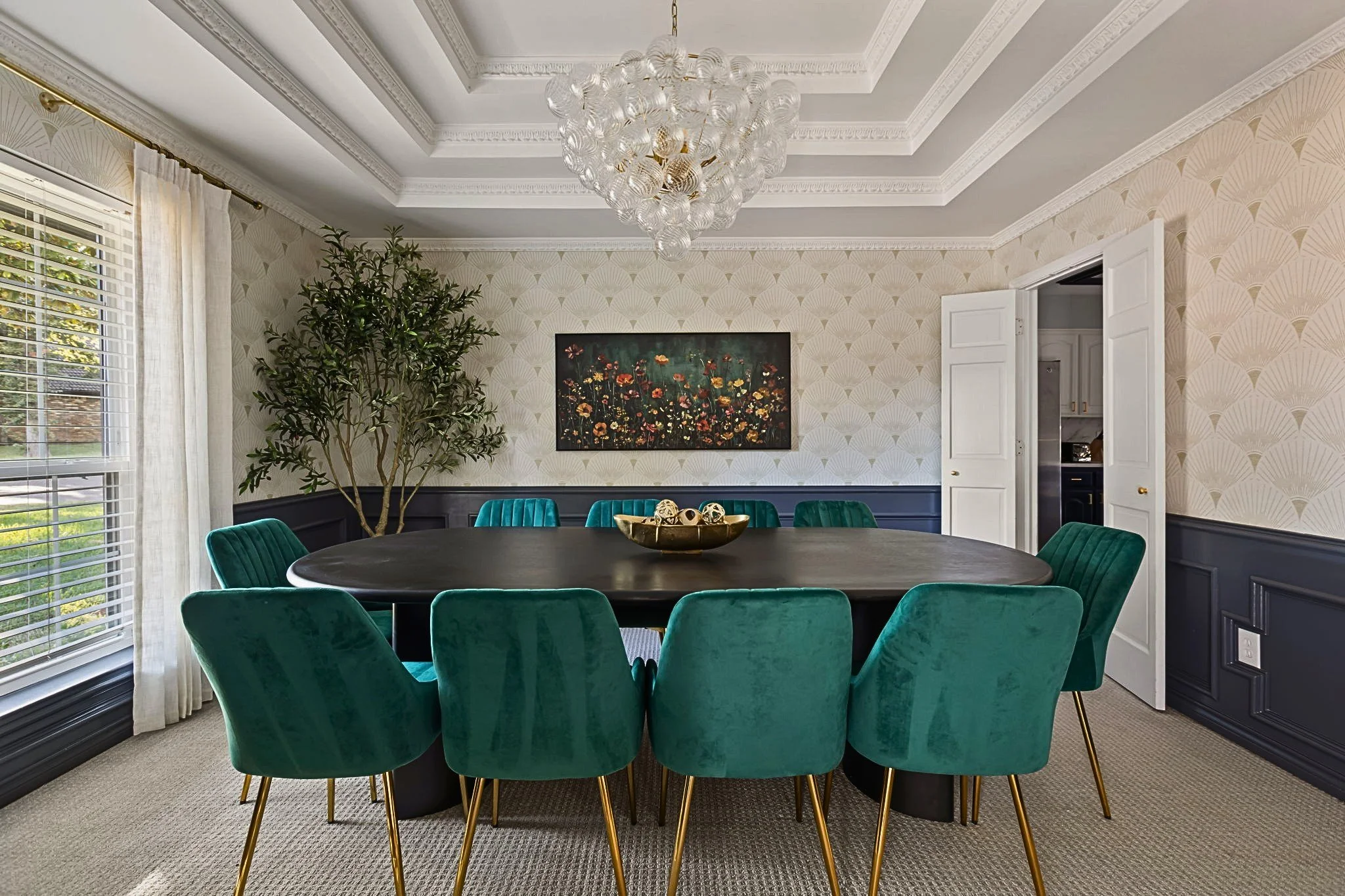 Modern dining room for a Dallas-Fort Worth area short-term rental designed by Kelsey Brown Interiors. The dining room features velvet, emerald green dining chairs, a black wooden dining table, and a globe chandelier. The room is decorated with wildfl
