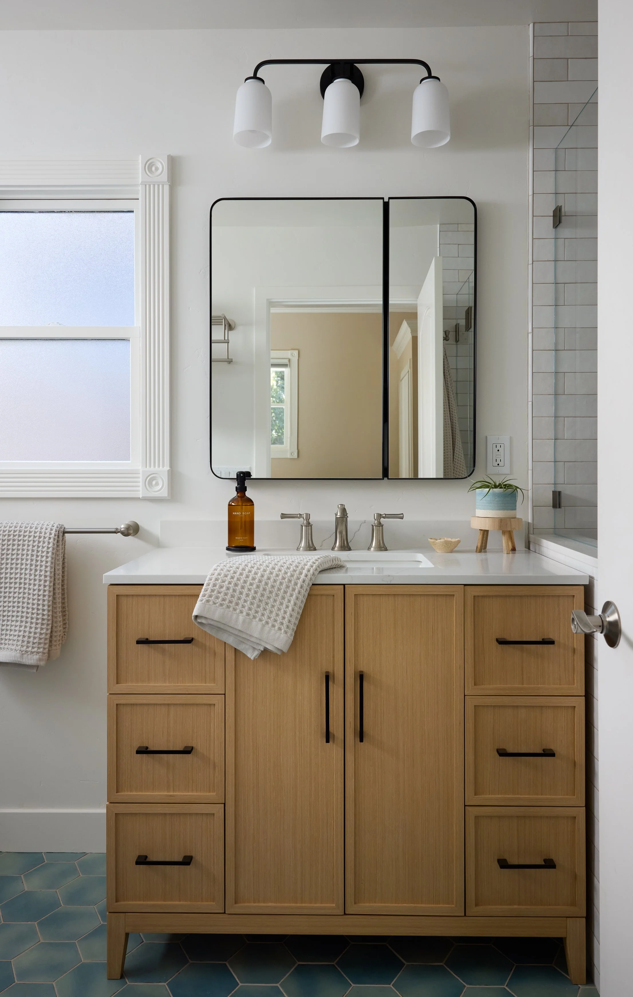 Transitional, coastal bathroom designed by Kelsey Brown Interiors in Aptos, California. Featuring a light oak vanity with black accents and a brushed nickel faucet. Blue green hexagon tiles as the bathroom flooring. 