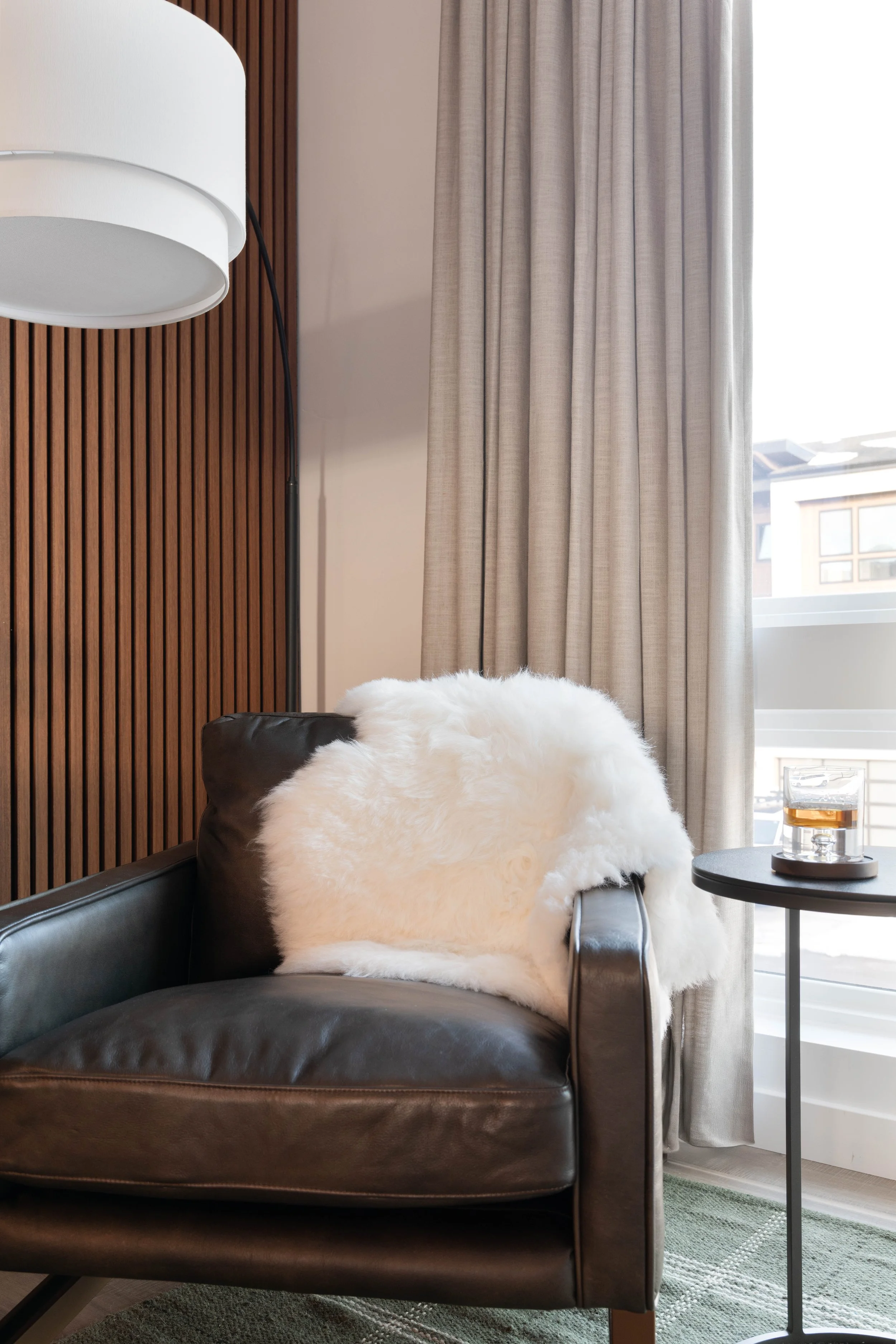 A black leather armchair with a white fluffy throw pillow, next to a small black round side table with a glass of amber-colored beverage, positioned near a window with draped light-colored curtains, a wooden wall accent, and a white modern floor lamp