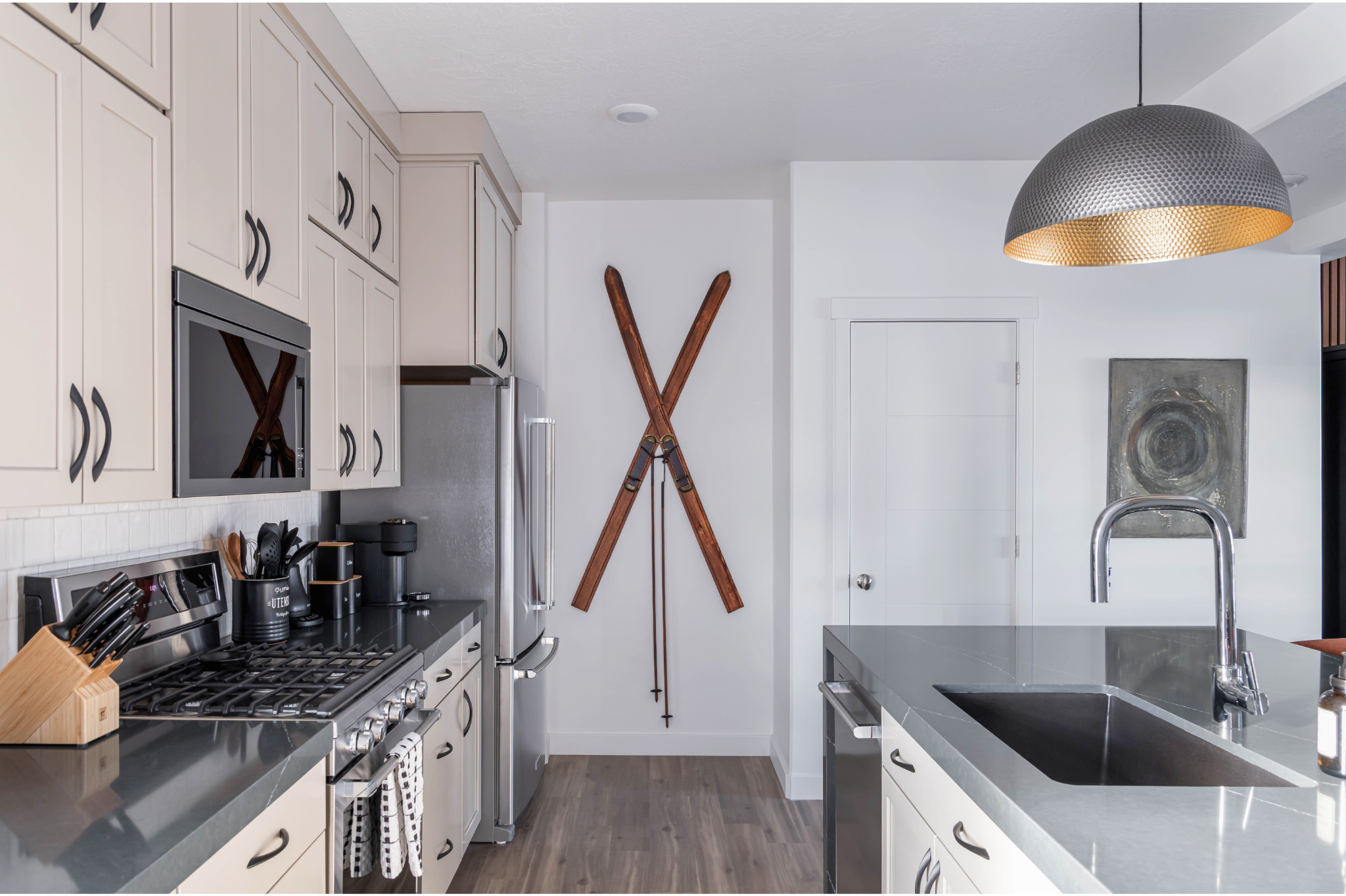 Kitchen with white cabinets, stainless steel appliances including an oven and refrigerator, a black countertop, and a gray island with a stainless steel sink. A set of vintage wooden skis is mounted on the white wall, and there is a modern hanging li