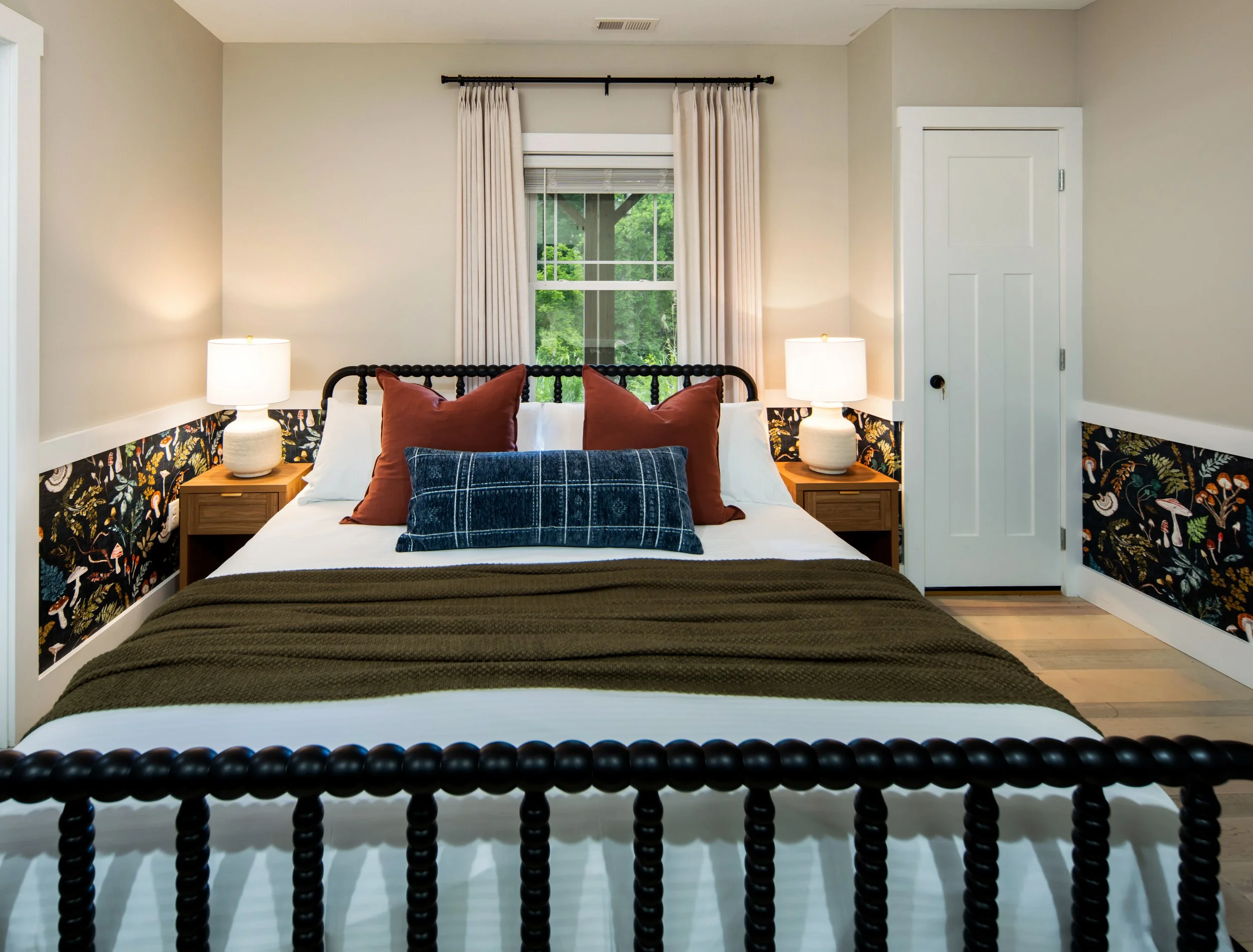 A short-term bedroom with a moody woodland inspired wallpaper, olive green knitted throw blanket, and rust colored throw pillows and blue lumbar pillow designed by Kelsey Brown Interiors in Asheville, North Carolina.