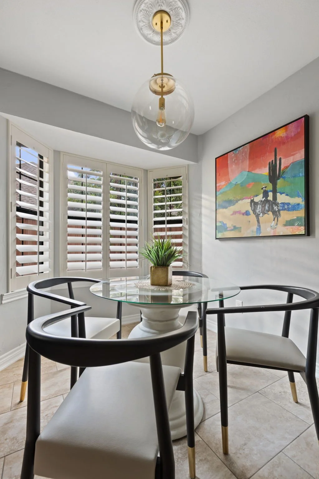 Kitchen nook in a Dallas-Fort Worth short-term rental featuring colorful artwork of a desert scene and a round gold and clear pendant over the glass dining table. Designed by Kelsey Brown Interiors.