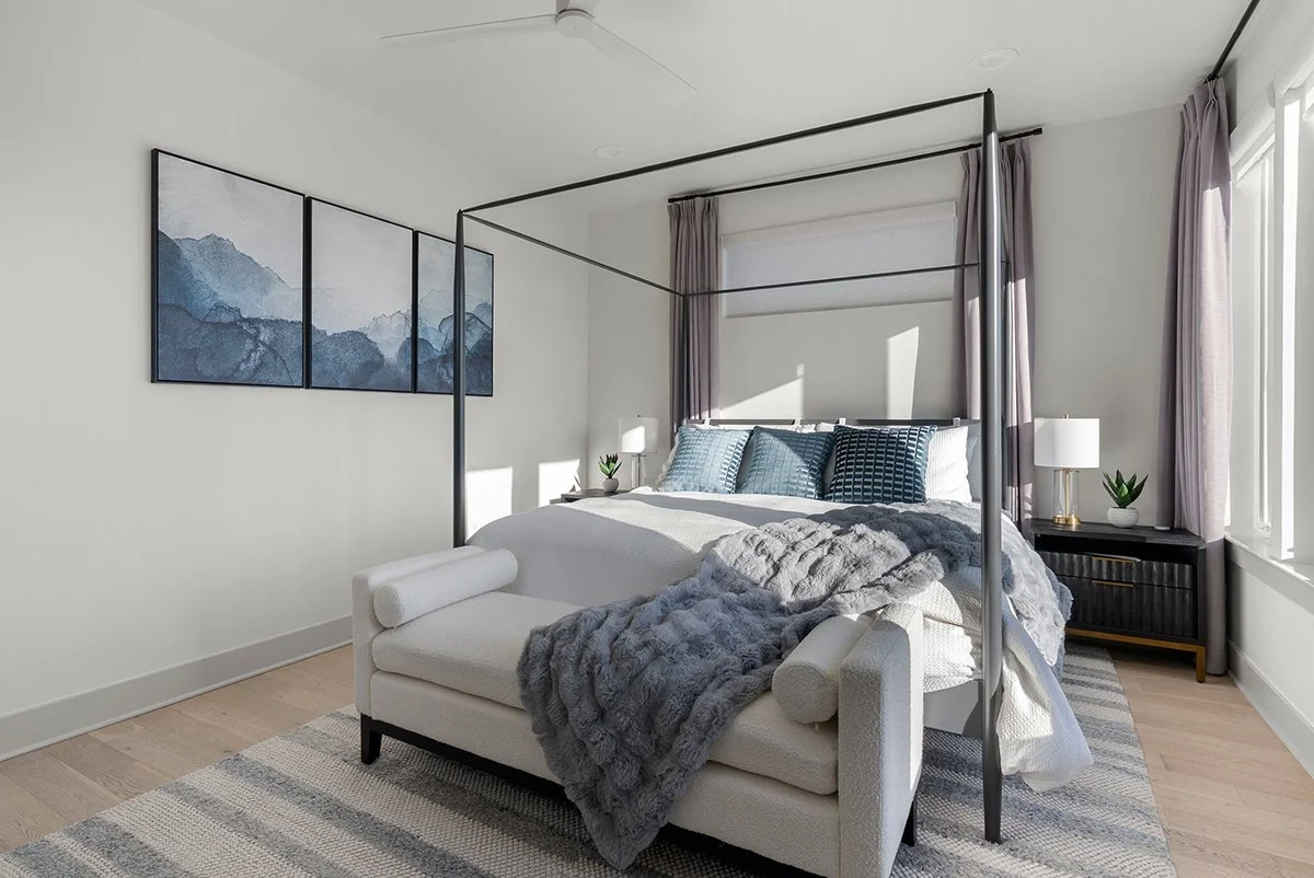 A modern bedroom with a white bed, gray throw blanket, blue pillows, three-part mountain-themed artwork on the wall, black four-poster bed frame, bedside tables with lamps, window with gray curtains, and a light-colored wooden floor.