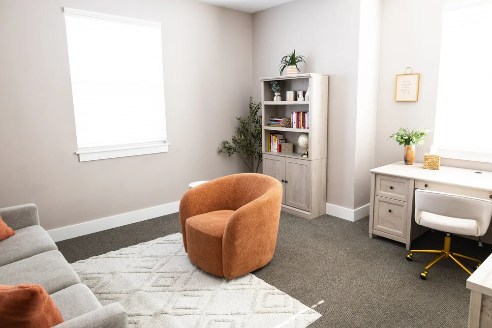 A therapy room at Healing Hearts Counseling in Murray, Utah, with a rust-colored swivel chair, a sofa, a bookshelf, plants and natural light.