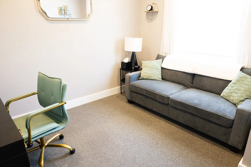 A therapy room at Healing Hearts Counseling in Murray, Utah, with a gray sofa, throw pillows, a lamp and a teal desk chair.