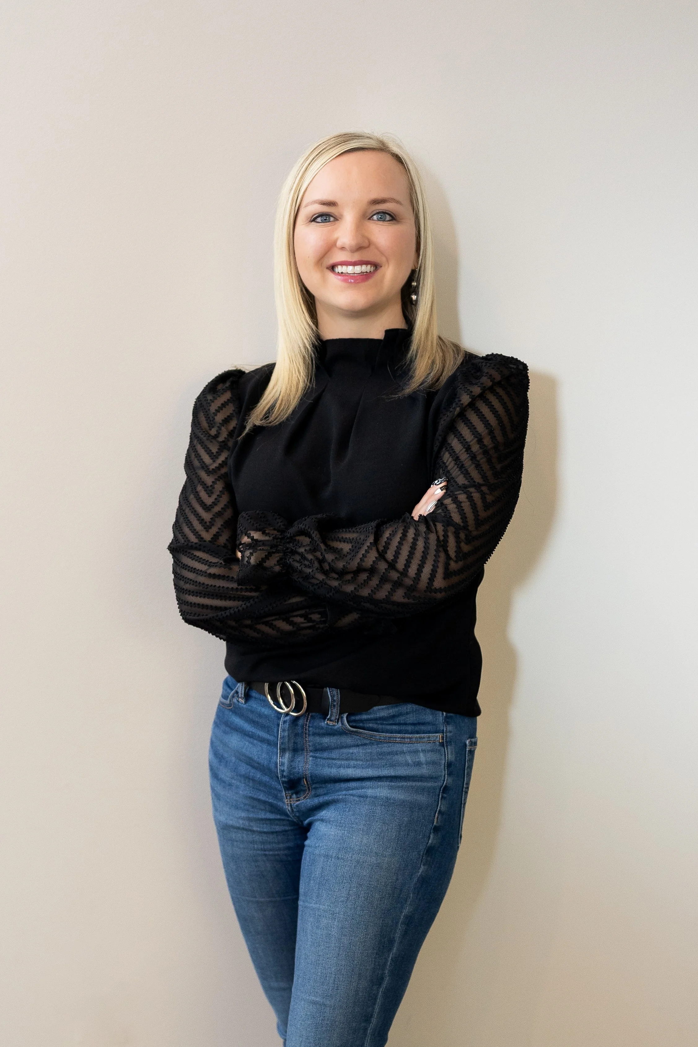 A woman with blonde hair wearing a sleeveless green top and black pants, standing against a plain beige wall, smiling at the camera.