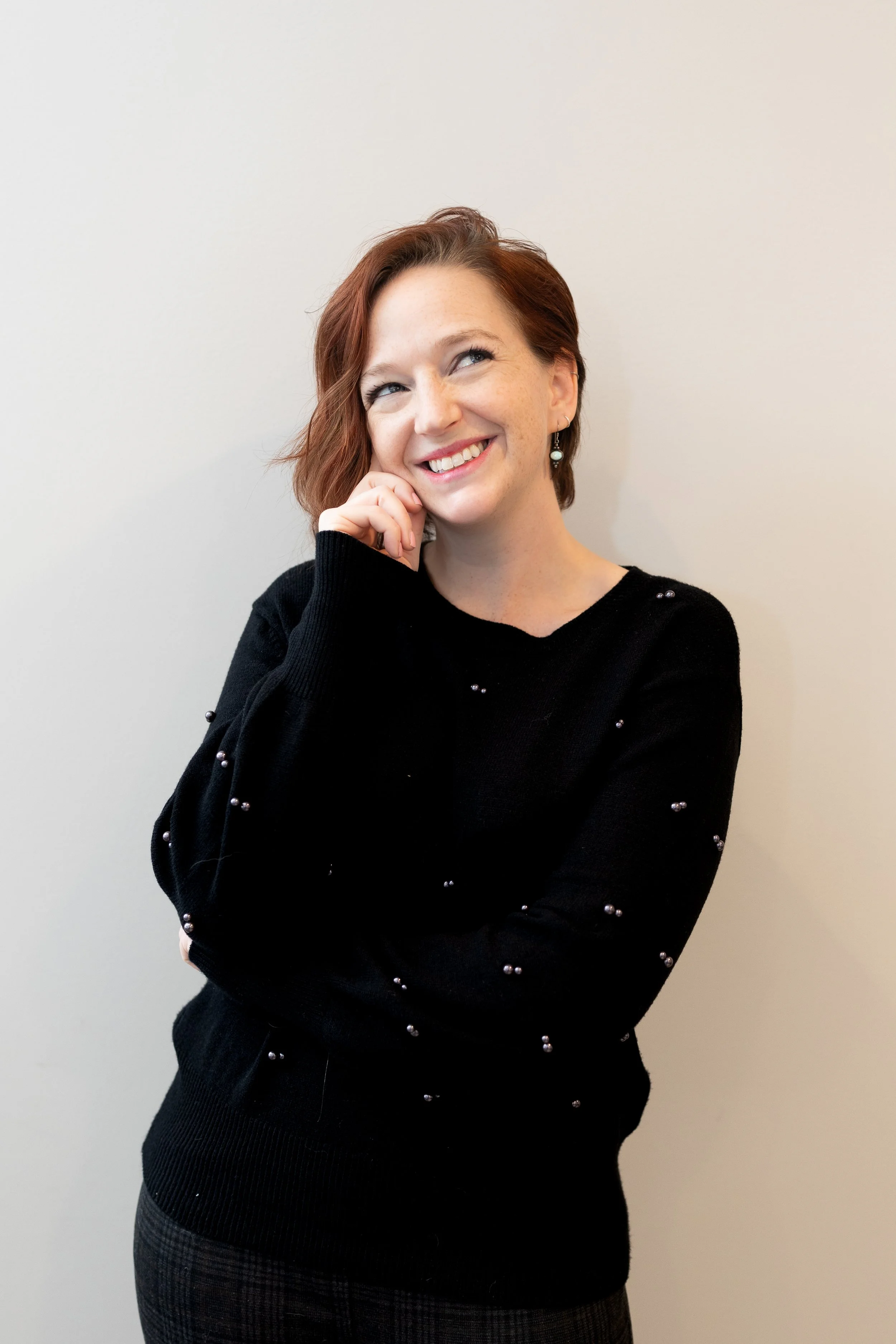 Portrait of a woman with short wavy brown hair and blue eyes, wearing a gray V-neck top and small earrings, smiling gently against a plain light background.