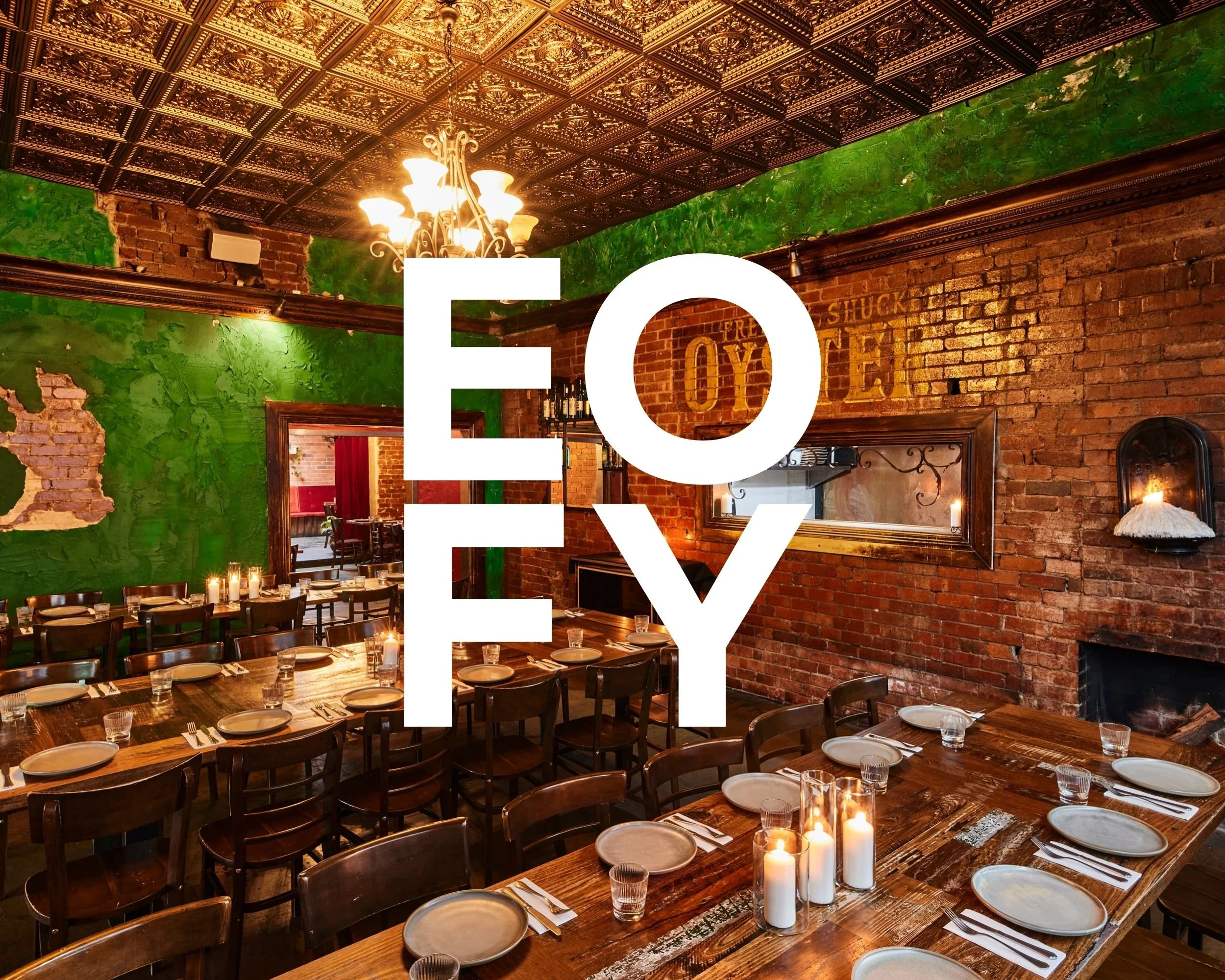 A cozy restaurant interior with exposed brick walls, green accents, a decorative tiled ceiling, and a chandelier. The dining area features wooden tables set with plates, glasses, and candles.