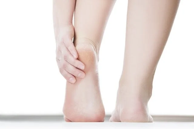 Une personne se tient le talon avec la main, montrant une douleur ou une gêne au niveau du talon.