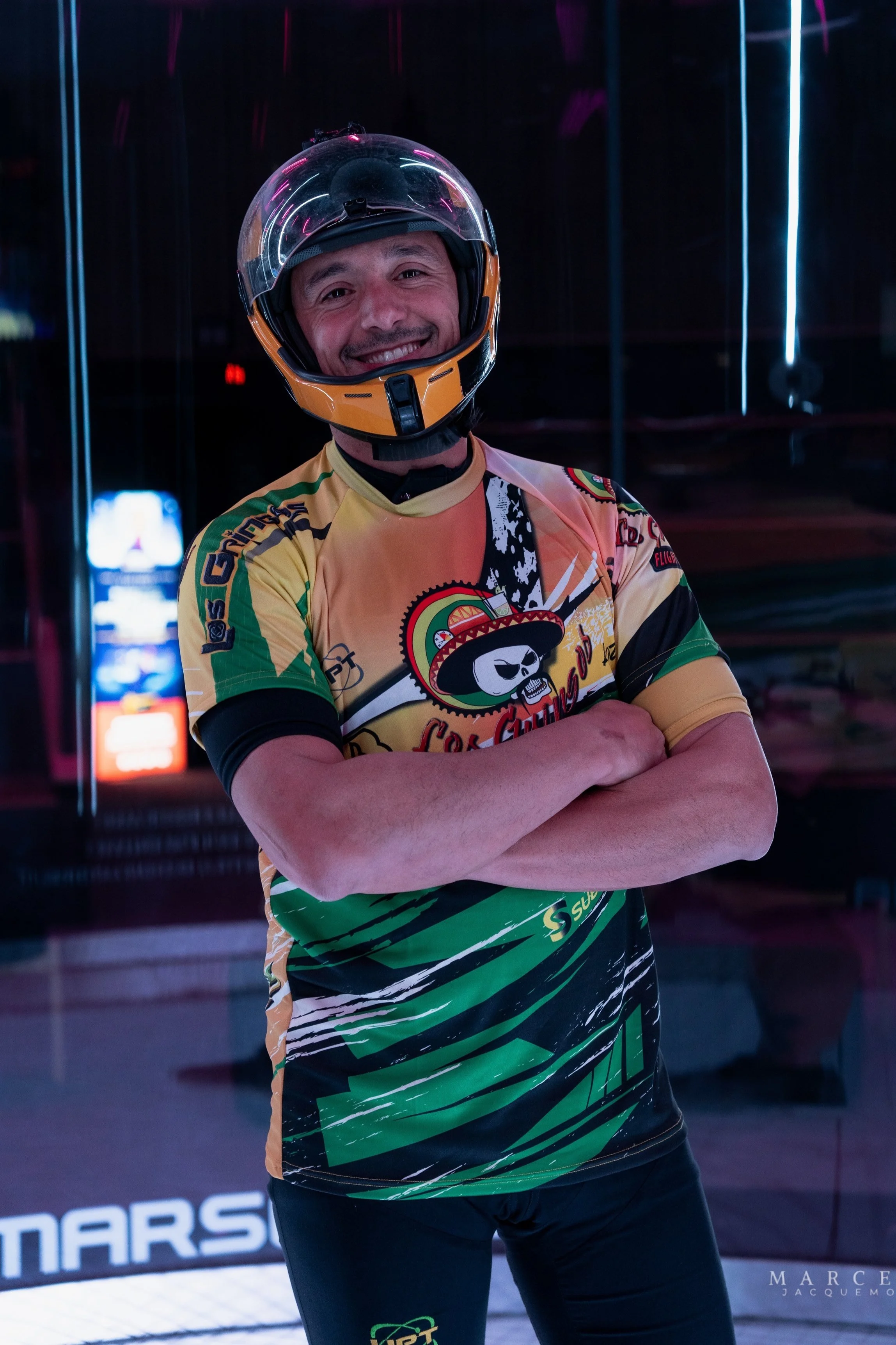 Un homme souriant portant un casque de moto et un maillot de sport coloré, avec ses bras croisés.
