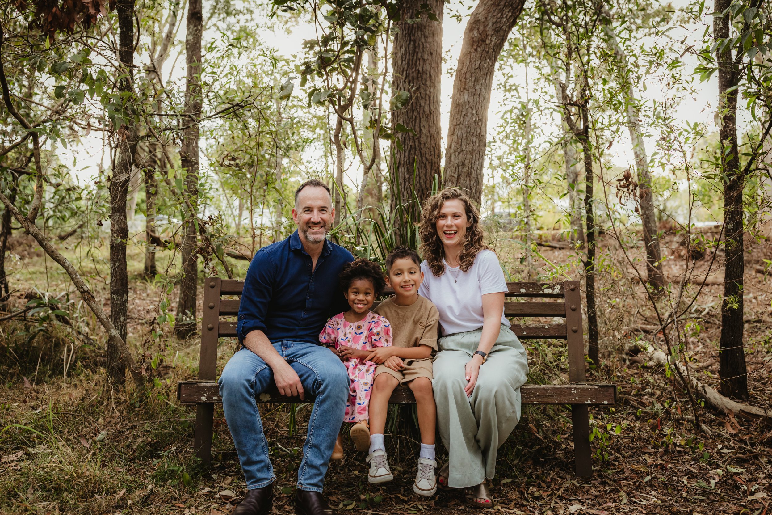 A family of four smiling outdoors. A man is holding a young boy with his arm around his neck. A woman wearing a hat is holding a toddler in a colorful sweater and pink dress. The background shows an open field and cloudy sky.