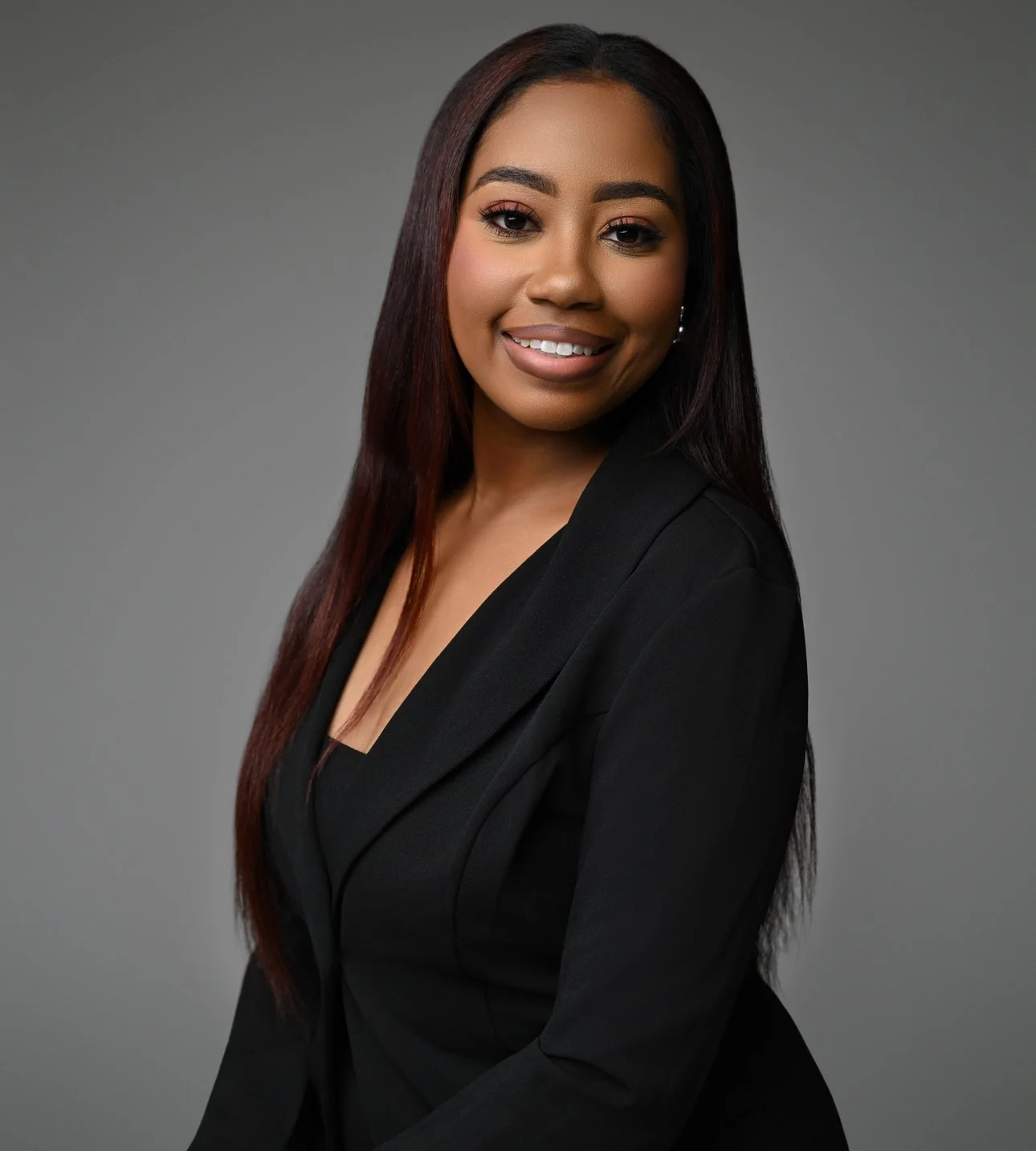 Professional portrait of a woman with long dark red hair, wearing a black blazer, smiling against a gray background.