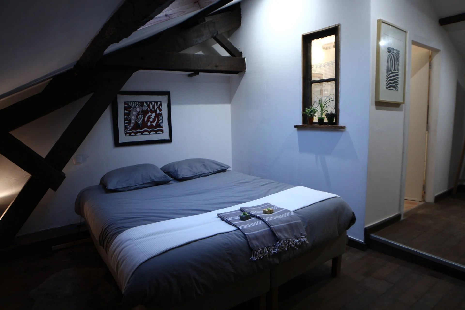 A cozy bedroom with a sloped ceiling, featuring a bed with gray bedding and two pillows. A small window with three potted plants on the sill, framed artwork hanging on the wall, and a wooden floor.