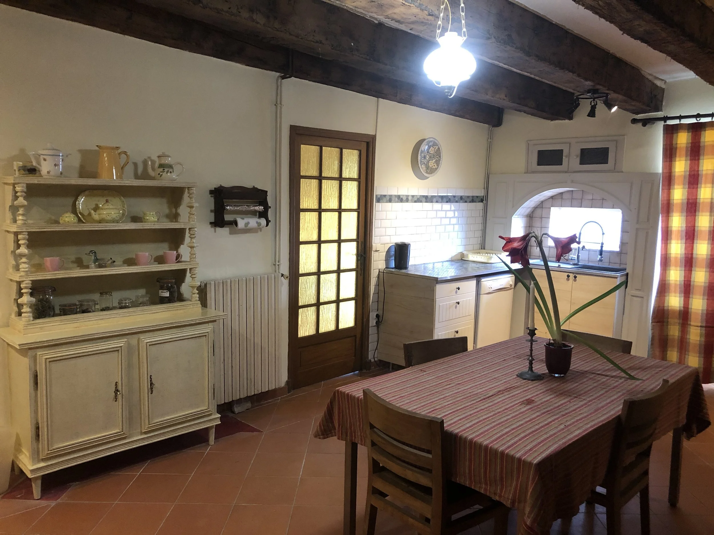Cozy rustic kitchen with wooden ceiling beams, a dining table with a striped tablecloth, a flower arrangement, and a vintage cabinet with decorative dishware, featuring a window with plaid curtains and a glass-paneled door.