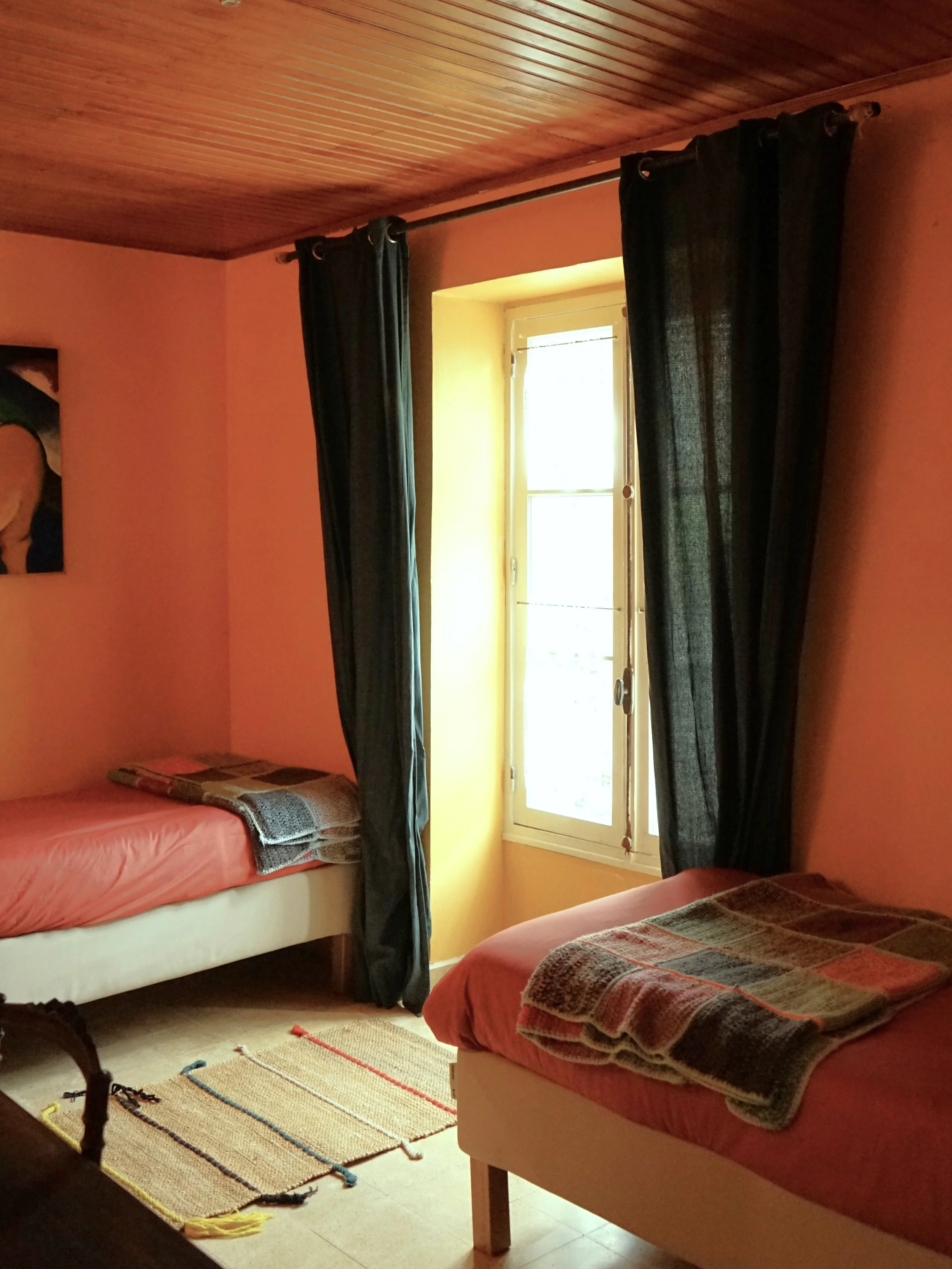A cozy bedroom with a yellow wall, a large bed with gray bedding and two pillows, a nightstand with a lamp, a colorful painting of a horse on the wall, and a window with dark curtains letting in natural light.
