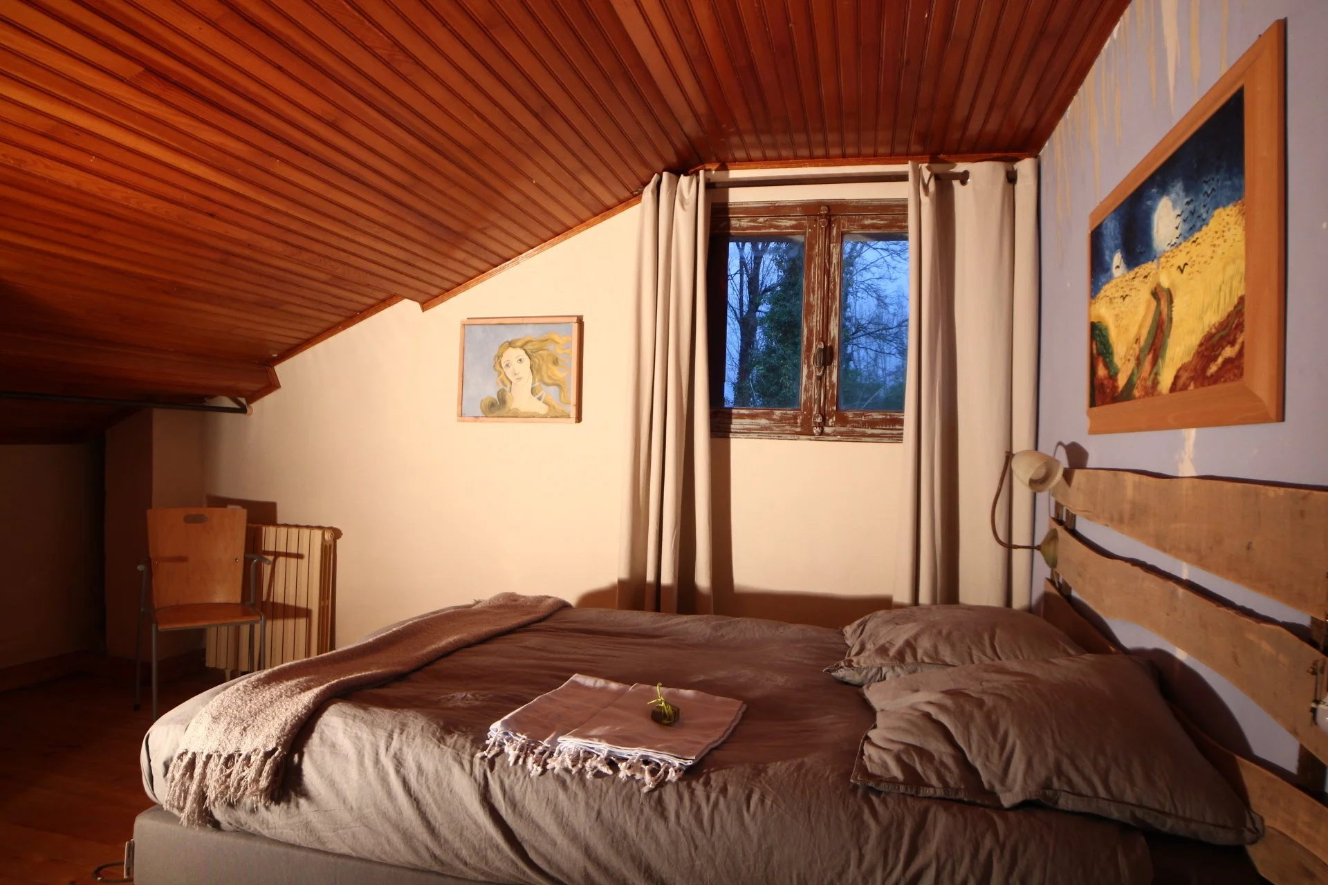 A cozy bedroom with a sloped wooden ceiling, light-colored walls, a window with beige curtains, and a bed with brown bedding and pillows. There are framed paintings on the walls and a small lamp attached to the headboard. A chair is against the wall near a radiator.