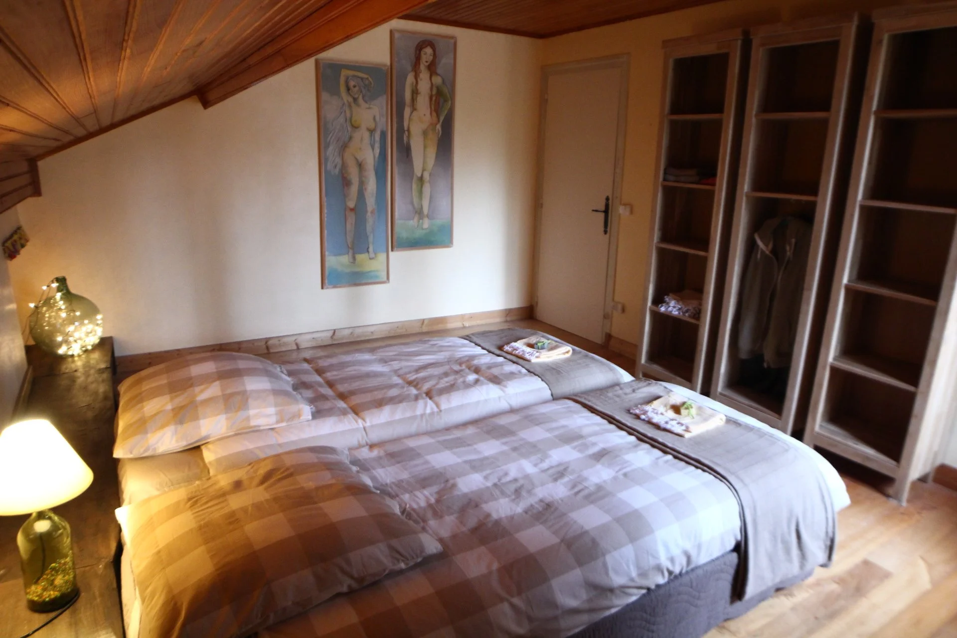 A cozy bedroom with two beds, wooden accents, wall artwork of two women, open wardrobe with clothes, and soft lighting.
