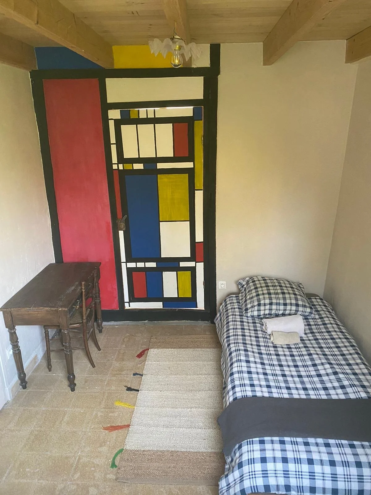 A small bedroom with a bed, a wooden desk, and a colorful stained-glass window. The bed has checkered bedding and towels on top. The floor is tiled with a rug and the ceiling has wooden beams.