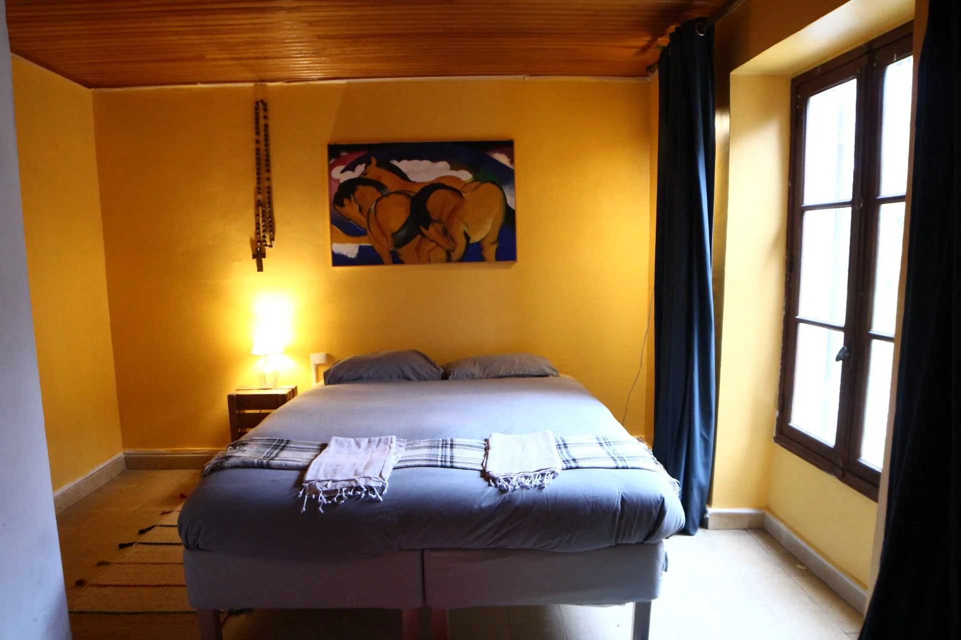 A bedroom with yellow walls, a bed with gray bedding, two gray pillows, and a checkered throw blanket. There is a bedside table with a lamp, framed artwork of horses on the wall, and a window with dark curtains.
