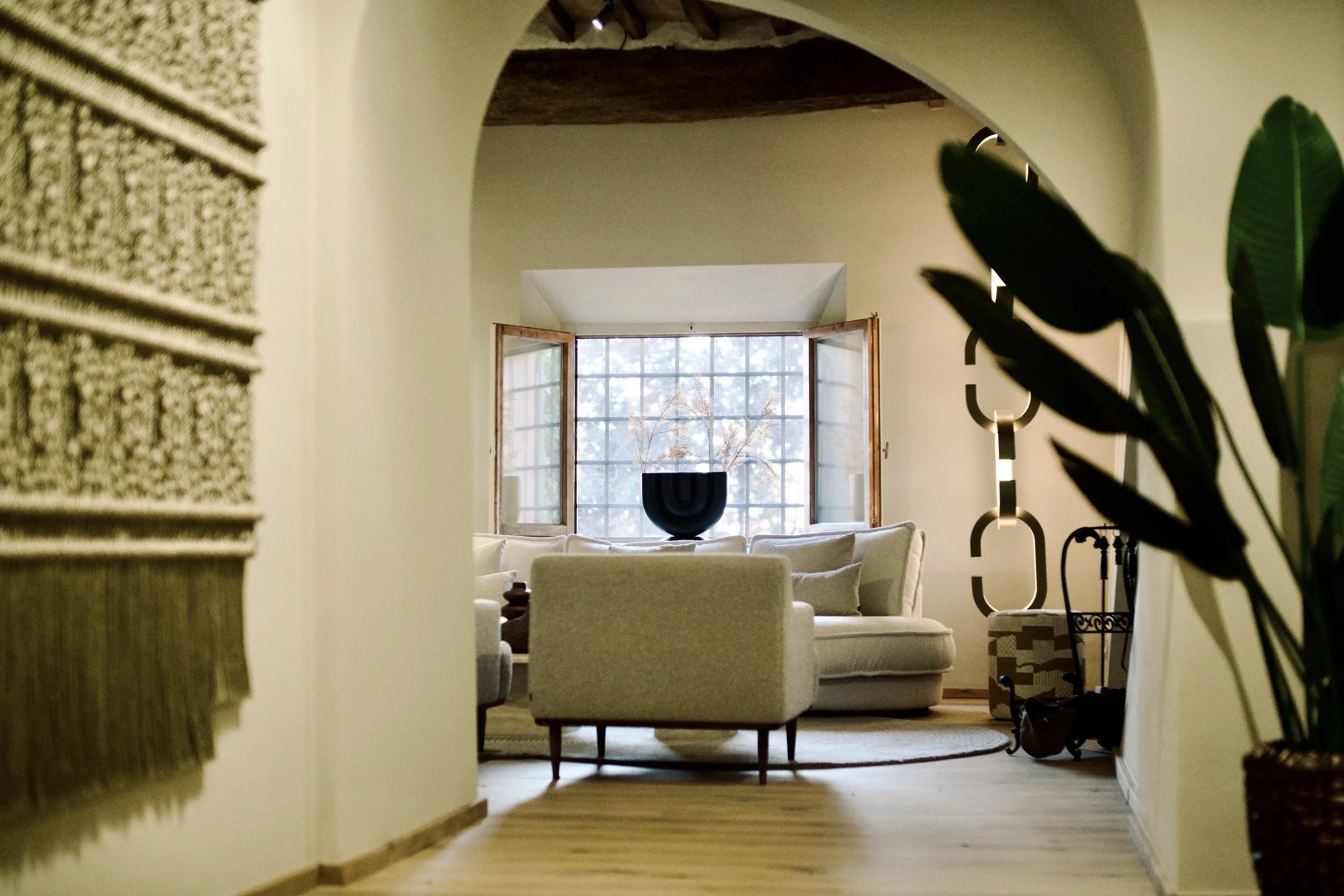 A cozy, well-lit living room with a small step-up area featuring a black patterned rug, a vintage wooden chair, and a guitar. Decor includes a large framed map on the wall, small sculptures, and a framed black-and-white photo. There are pink upholstered chairs in the foreground and a window with grid panes in the background, letting in natural light.