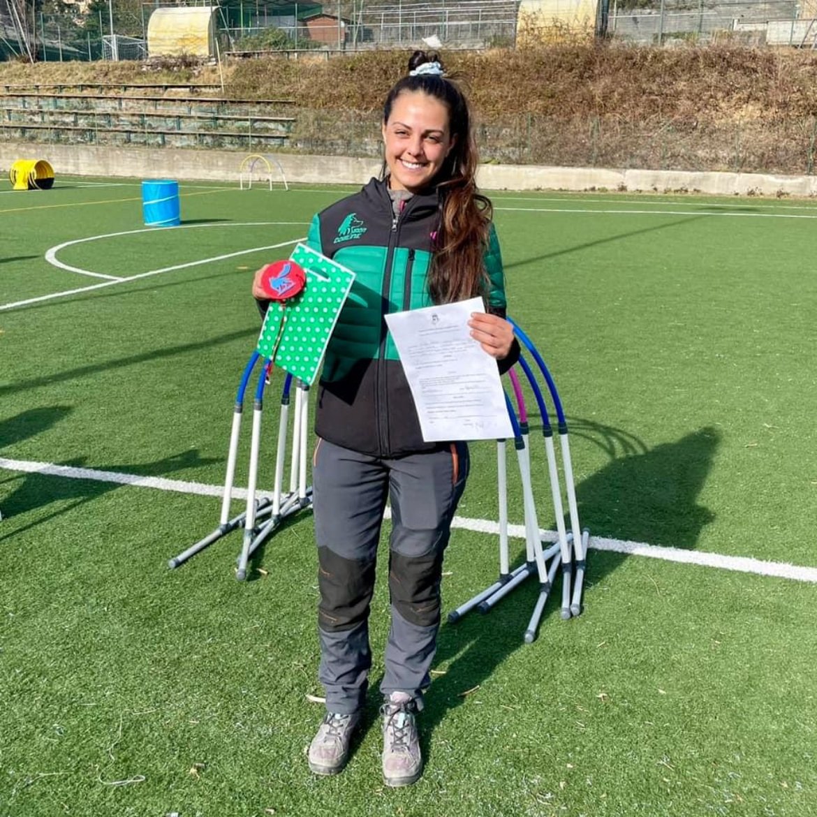 Una donna sorridente che tiene un foglio e alcuni oggetti mentre si trova su un campo sportivo.