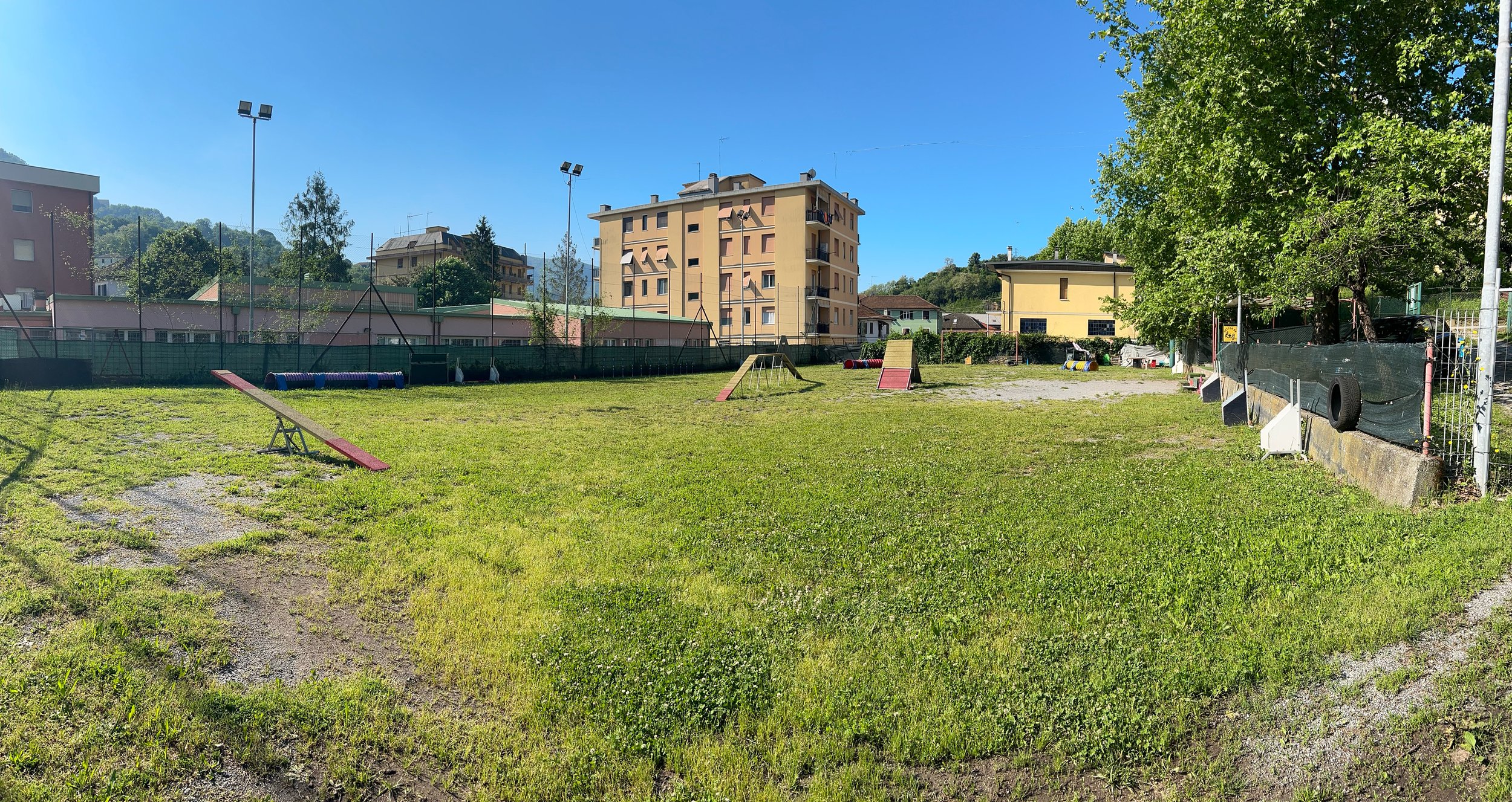 campo di addestramento per cani con ostacoli come rampe e tunnel, recinto con rete e alcuni edifici e alberi sullo sfondo.