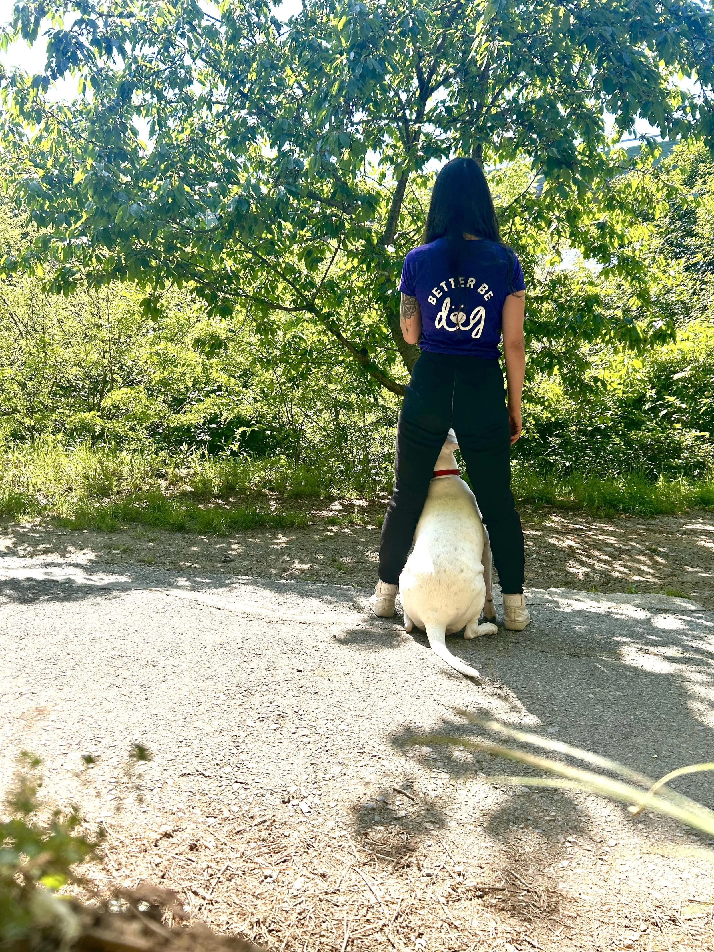 Una donna con pantaloni neri e una maglietta blu che dice 'Better Be a Dog' davanti a un albero, con un cane seduto tra le sue gambe, in un'area all'aperto con molte foglie verdi.