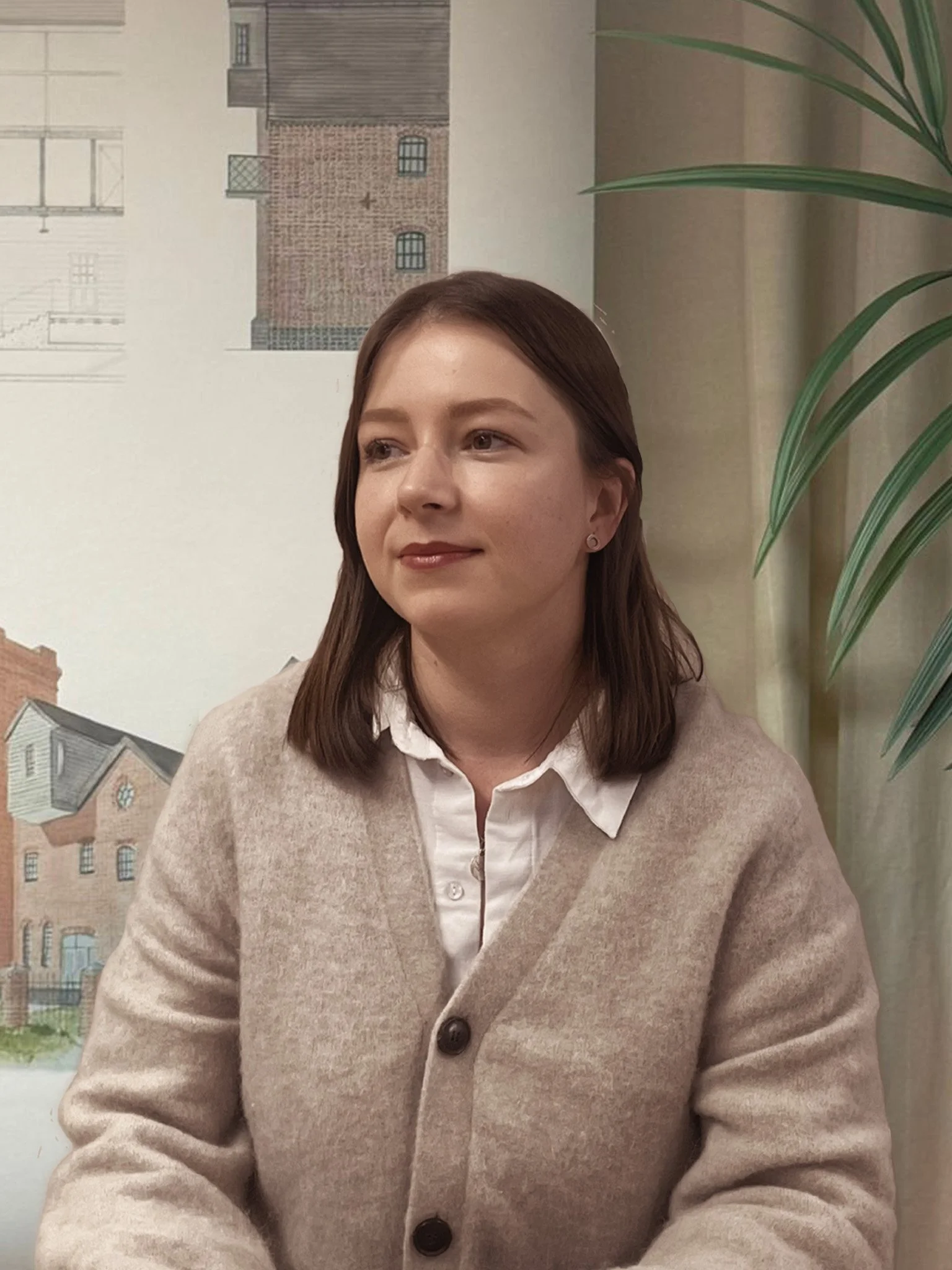 Julia from the 24/Three team in a grey cardigan posing in front of a green plant and a watercolour building sketch