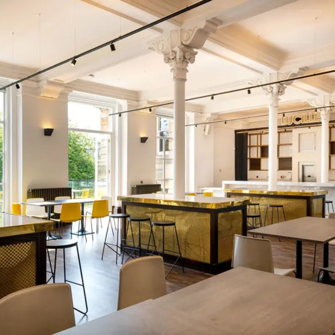 Modern cafe interior with high ceilings, large windows, and stylish furniture. Features include white pillars, wooden floors, tables with yellow and white chairs, and a bar with golden accents.