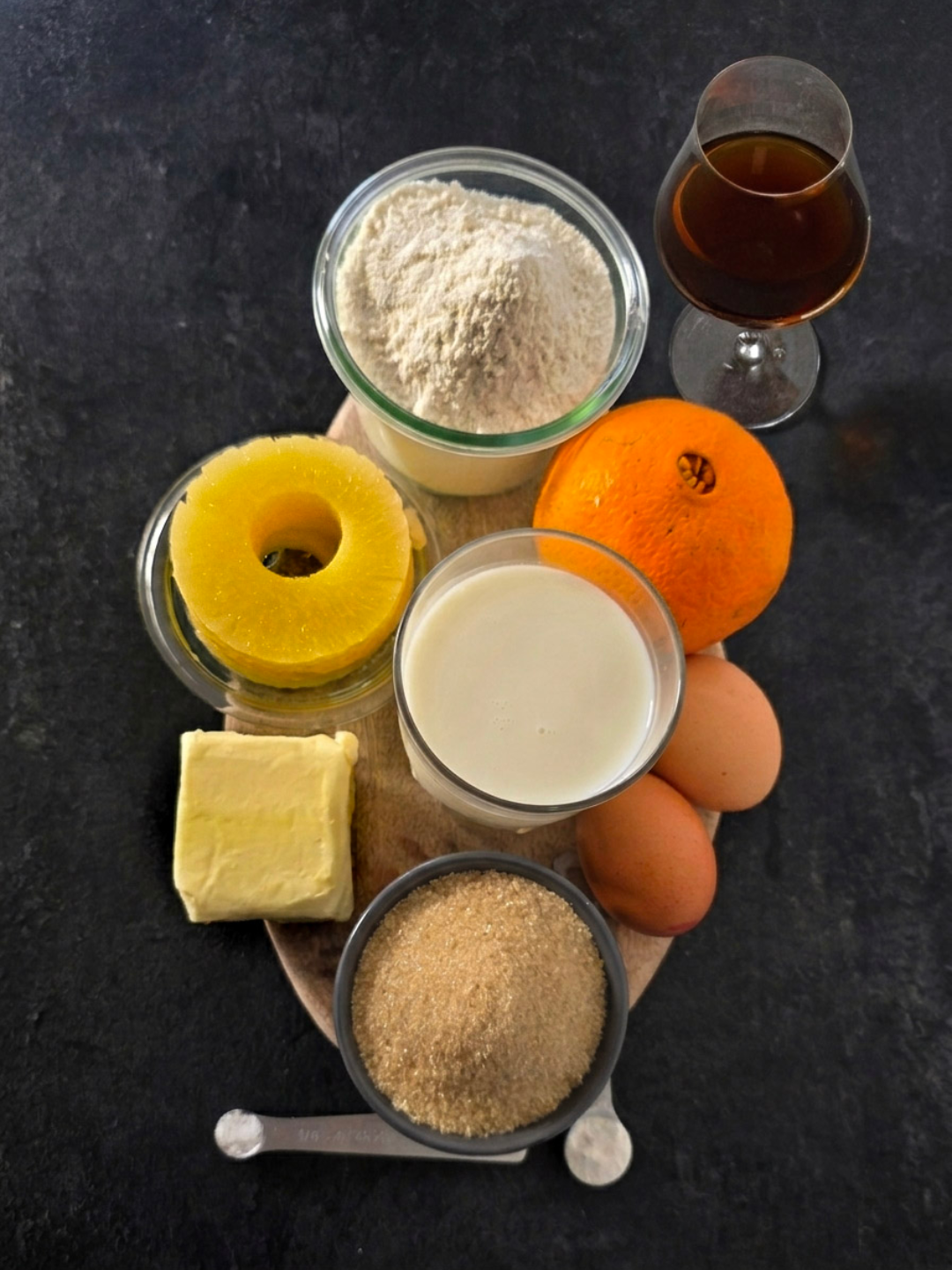 Flat lay of baking ingredients for Mulongo's Pineapple Upside-Down Cake on a dark slate surface — T55 flour, brown sugar, butter, whole milk, eggs, fresh orange and pineapple alongside a glass of Rivesaltes Ambré 1985.
