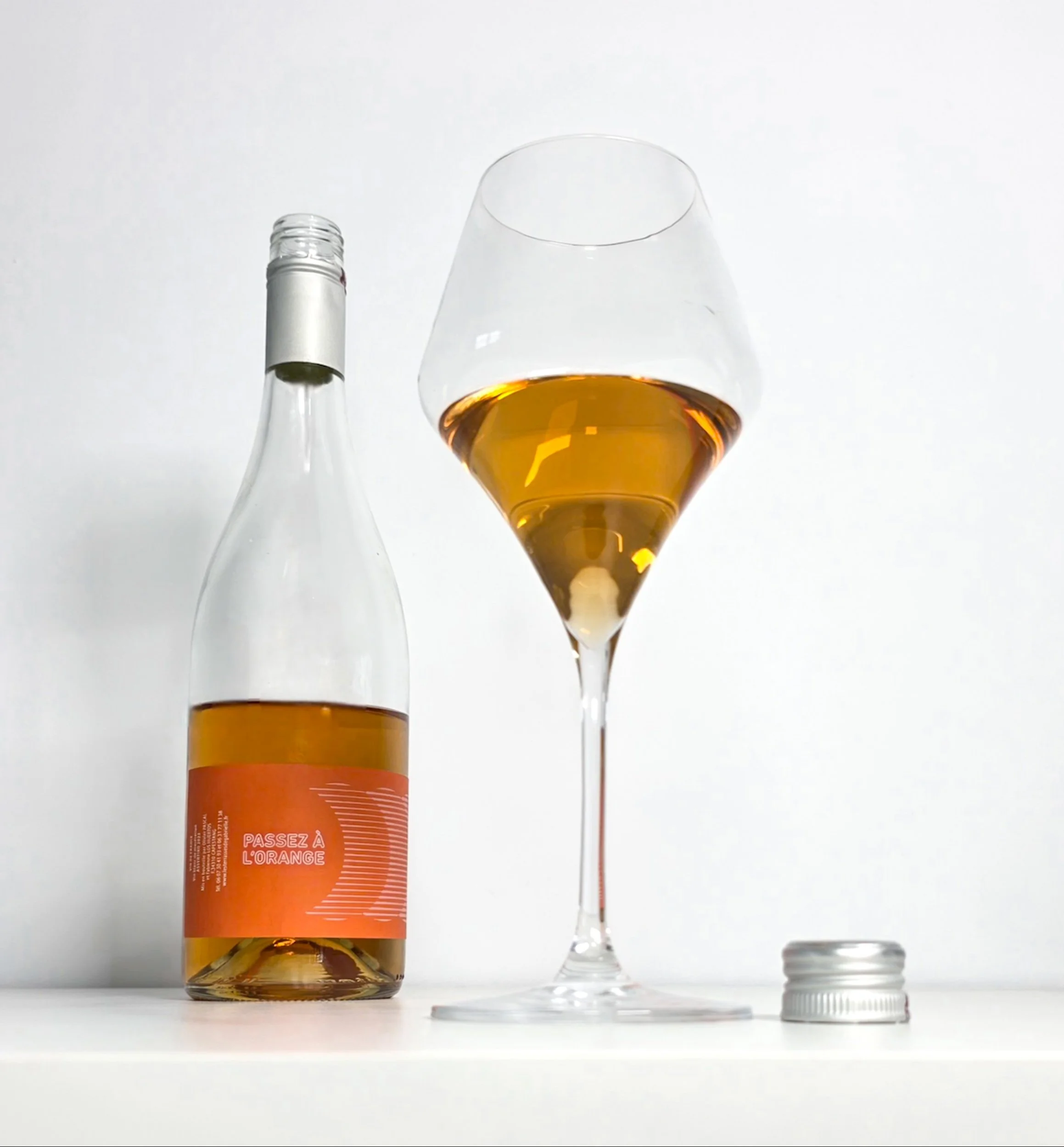 Glass and bottle of Passez à l'Orange Assyrtiko 2023 orange wine against a dark slate backdrop, showing the wine's distinctive amber colour. Vin de France by Pascal Olivier and Fabienne Los Huertos.