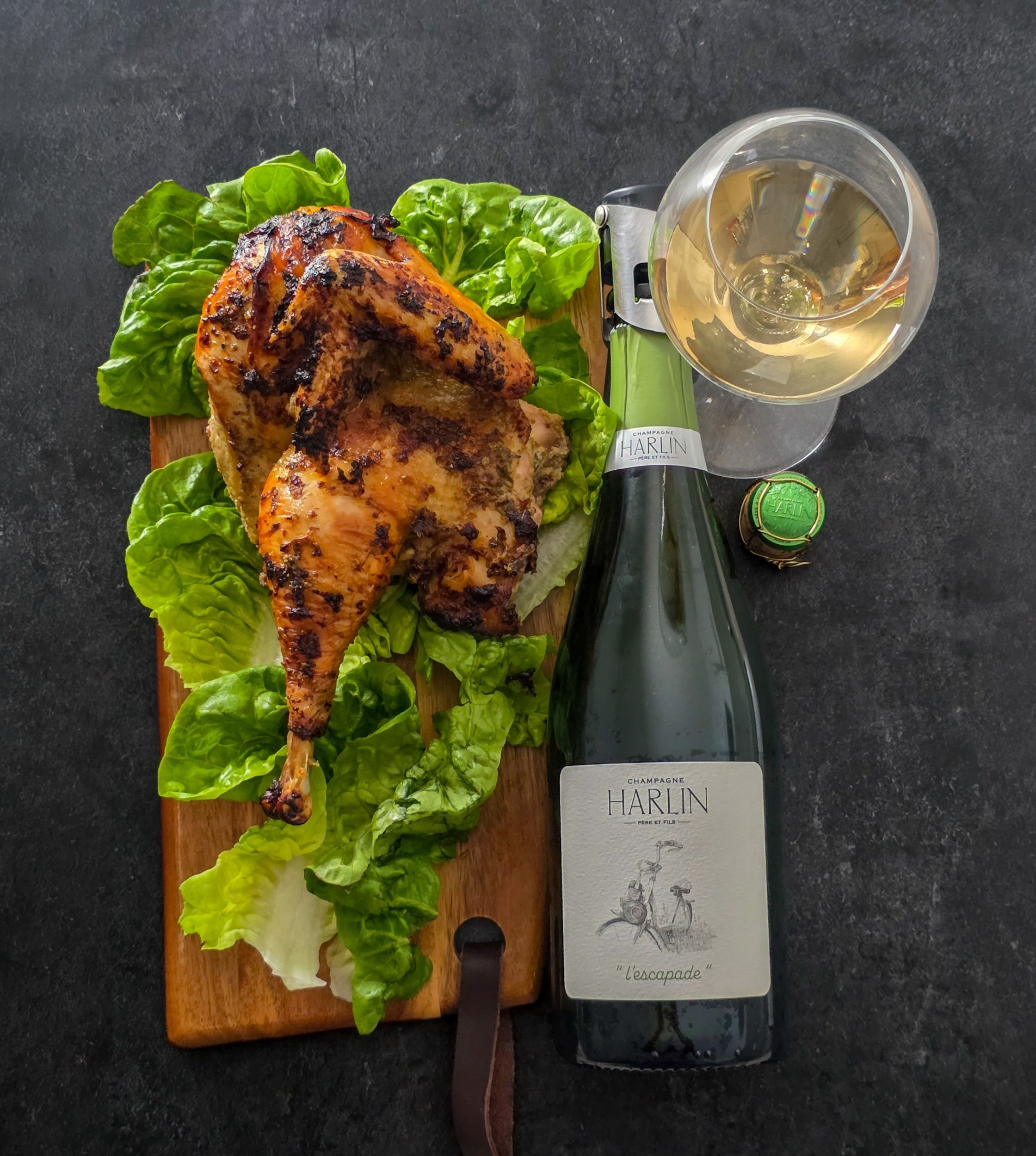 Overhead view of a roast half chicken on lettuce beside a bottle of Harlin L’Escapade Champagne, a glass and a green cap.