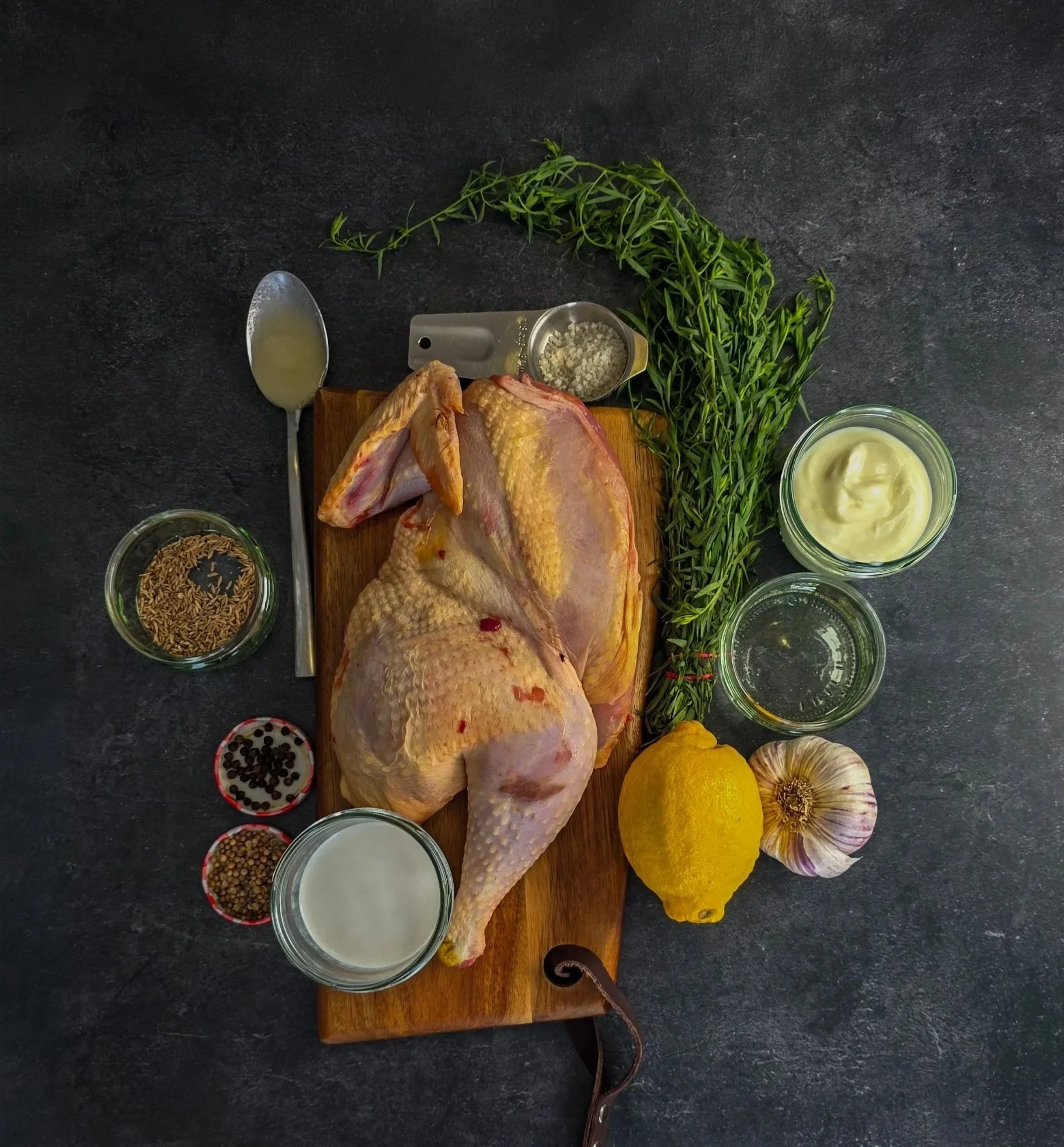 Flat lay of a raw half chicken, tarragon, lemon, garlic, yoghurt, coconut milk and spices on dark slate.