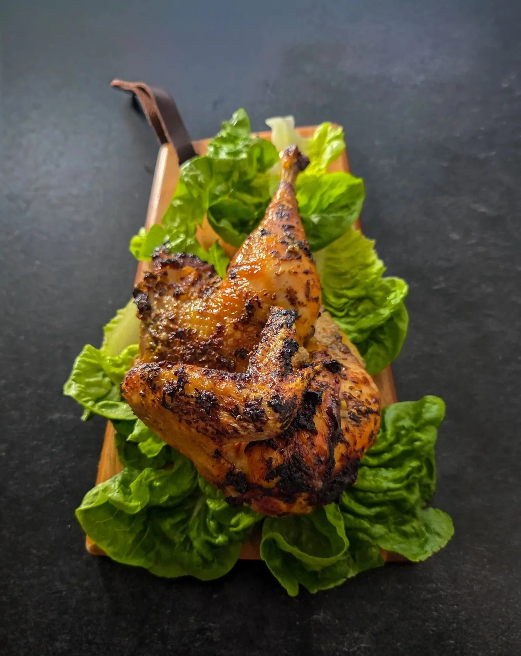 Golden roast half chicken with spiced skin on fresh lettuce on a wooden board, seen from above.