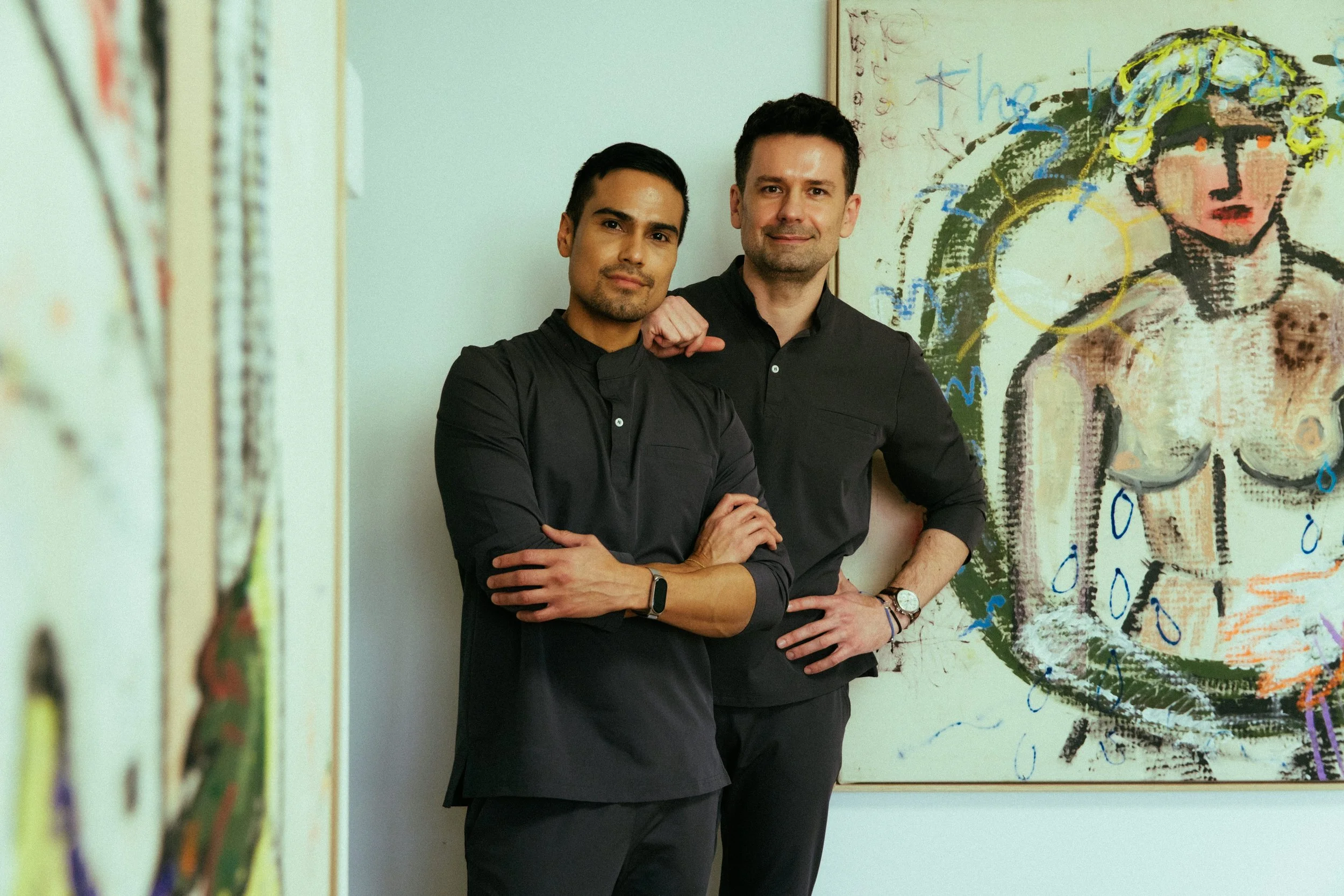 Two men standing in front of colorful artwork, one with arms crossed and the other with hand on hip, posing confidently.