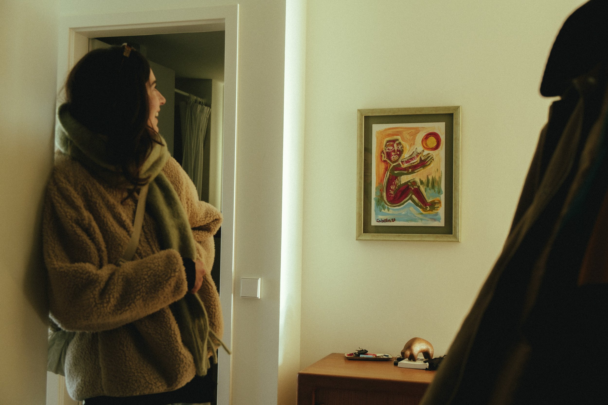 A woman wearing a fuzzy jacket, holding a scarf, leaning against a wall, looking at a man whose side is partially visible, standing indoors in a room. There is a framed colorful abstract painting on the wall and a wooden table with small objects on i
