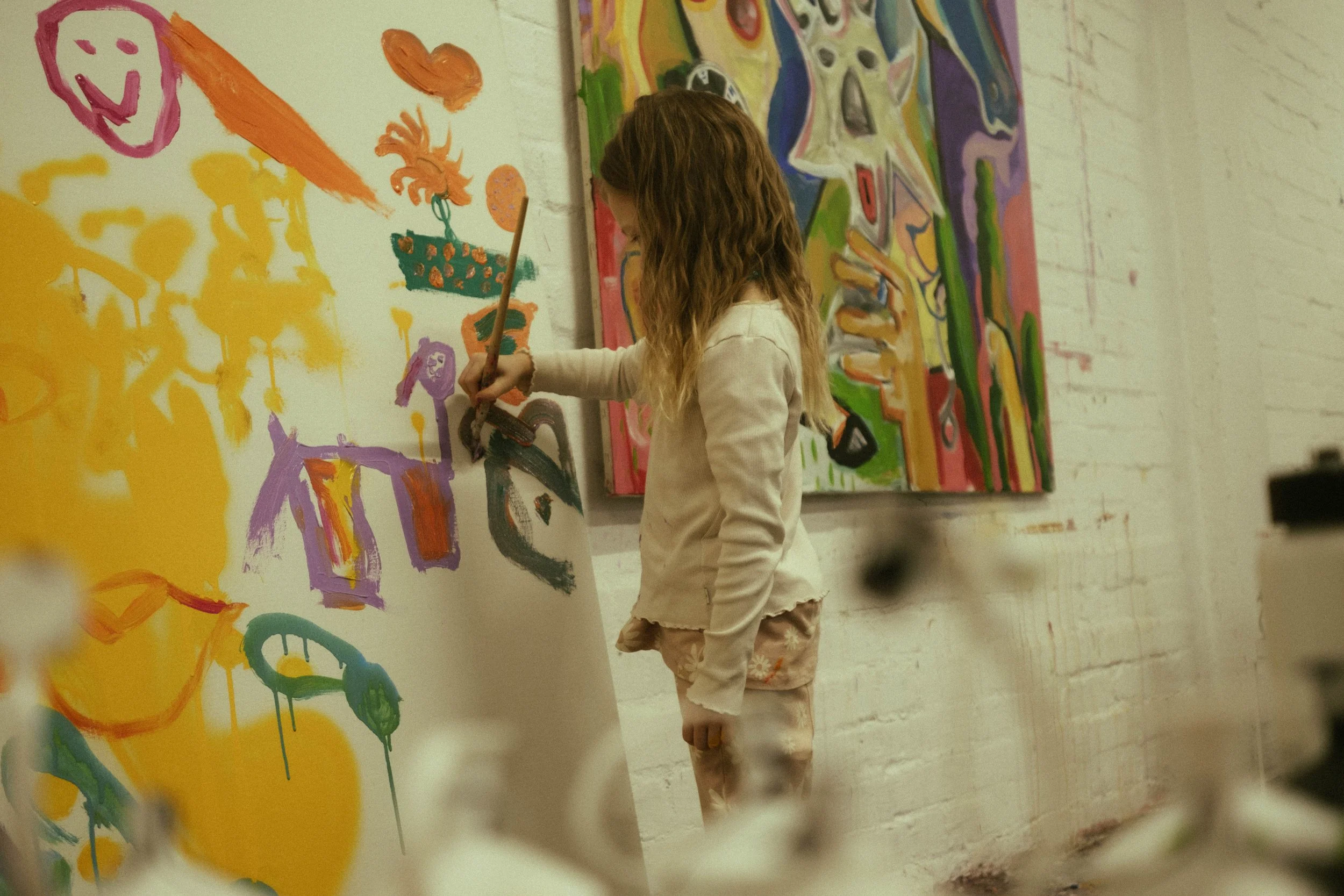 A girl with long curly hair wearing a beige long-sleeve shirt and matching shorts, painting on a large canvas. The art on the canvas includes colorful, abstract shapes and lines, with a visible smiling face drawn with pink paint on the wall behind her in an art studio or classroom.