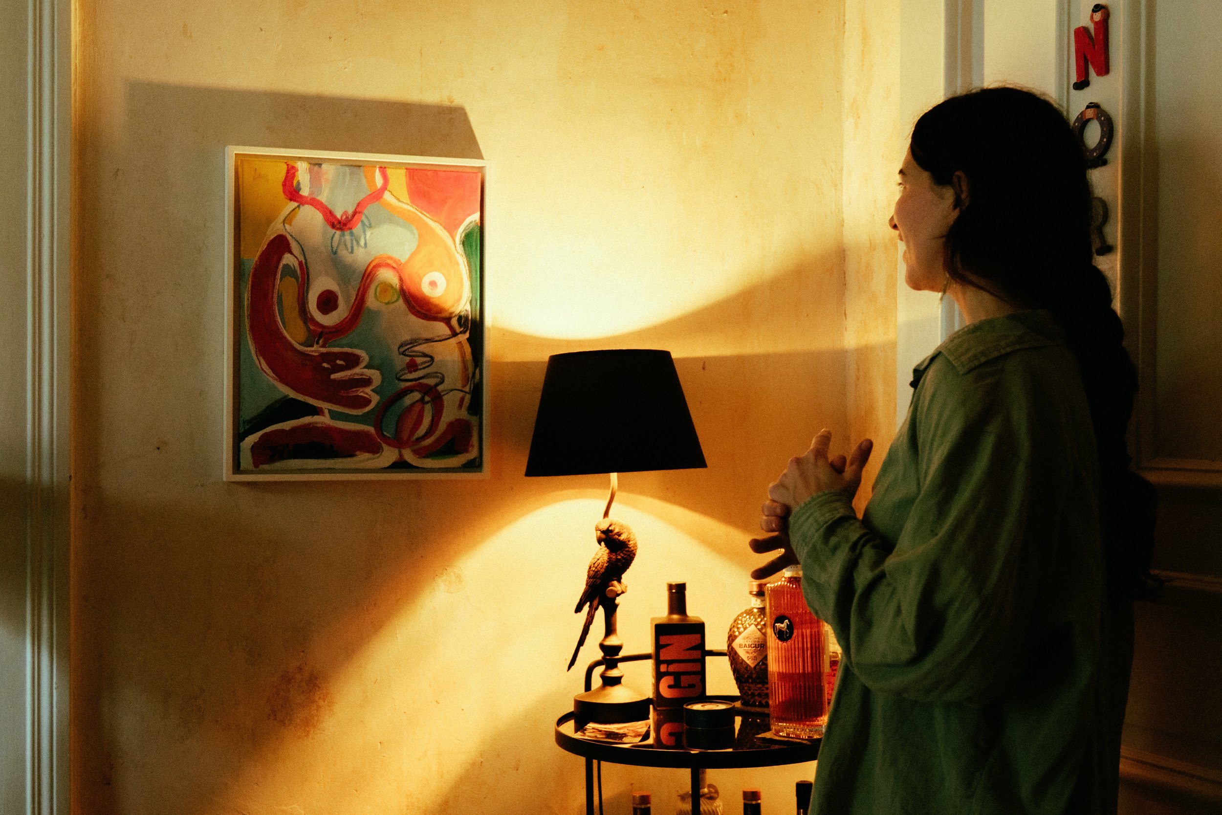 A woman standing in a dimly lit room, looking at a colorful abstract painting on the wall, illuminated by a black lampshade on a small table with various bottles and decorative items.