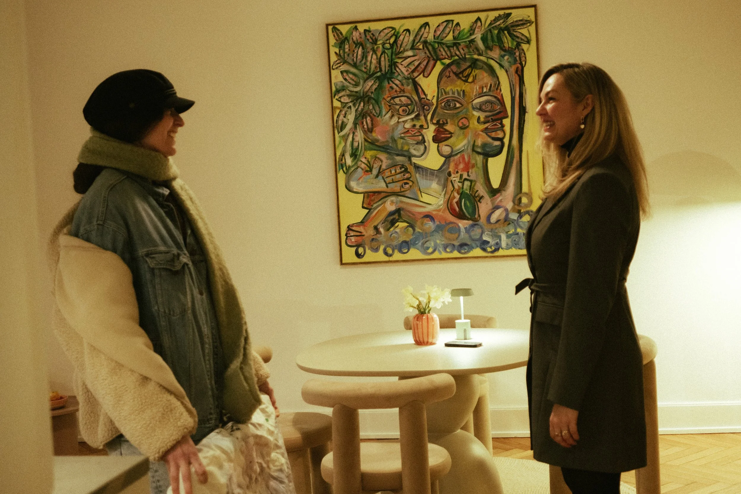 Two women standing and talking in a warmly lit room with a colorful abstract painting on the wall in the background, a small round table with a flower vase and a lamp.