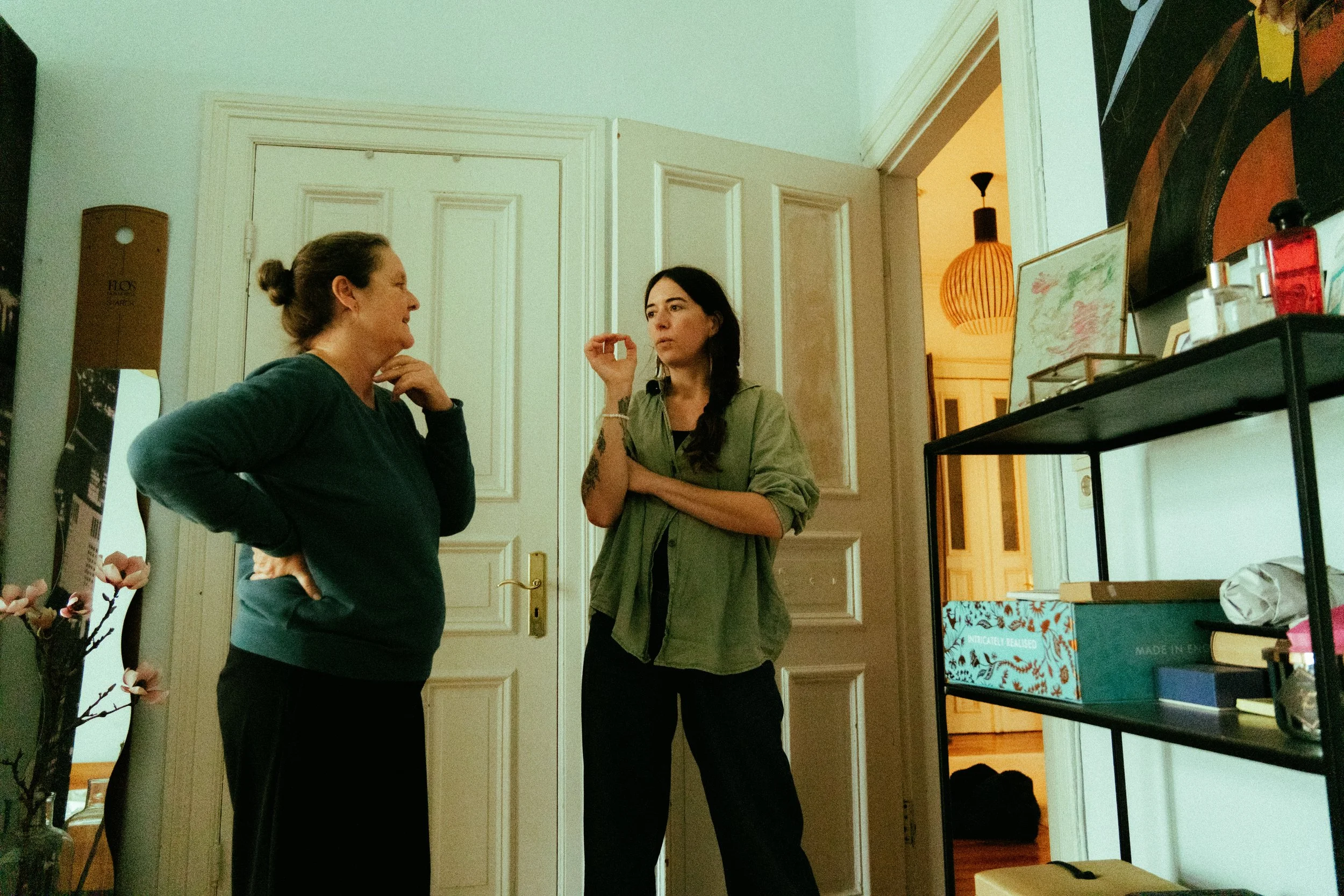 Two women standing and talking in a room with an open door, decorated with paintings and shelves with various items.