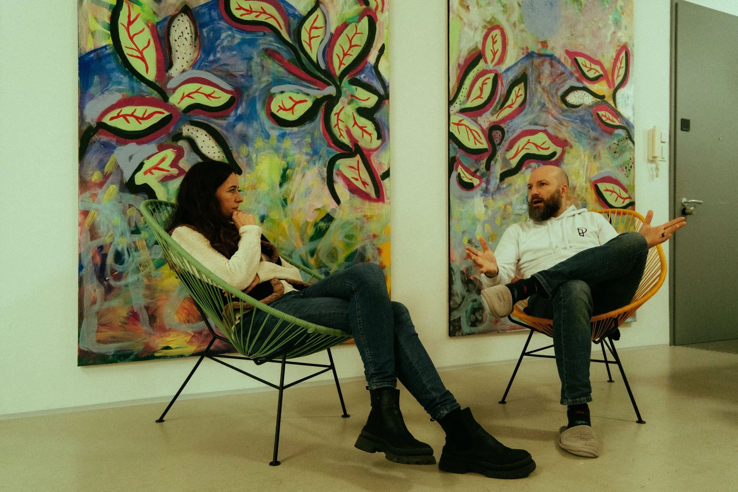 Two people sitting in colorful chairs having a conversation in front of vibrant abstract paintings with leaves on the wall behind them.